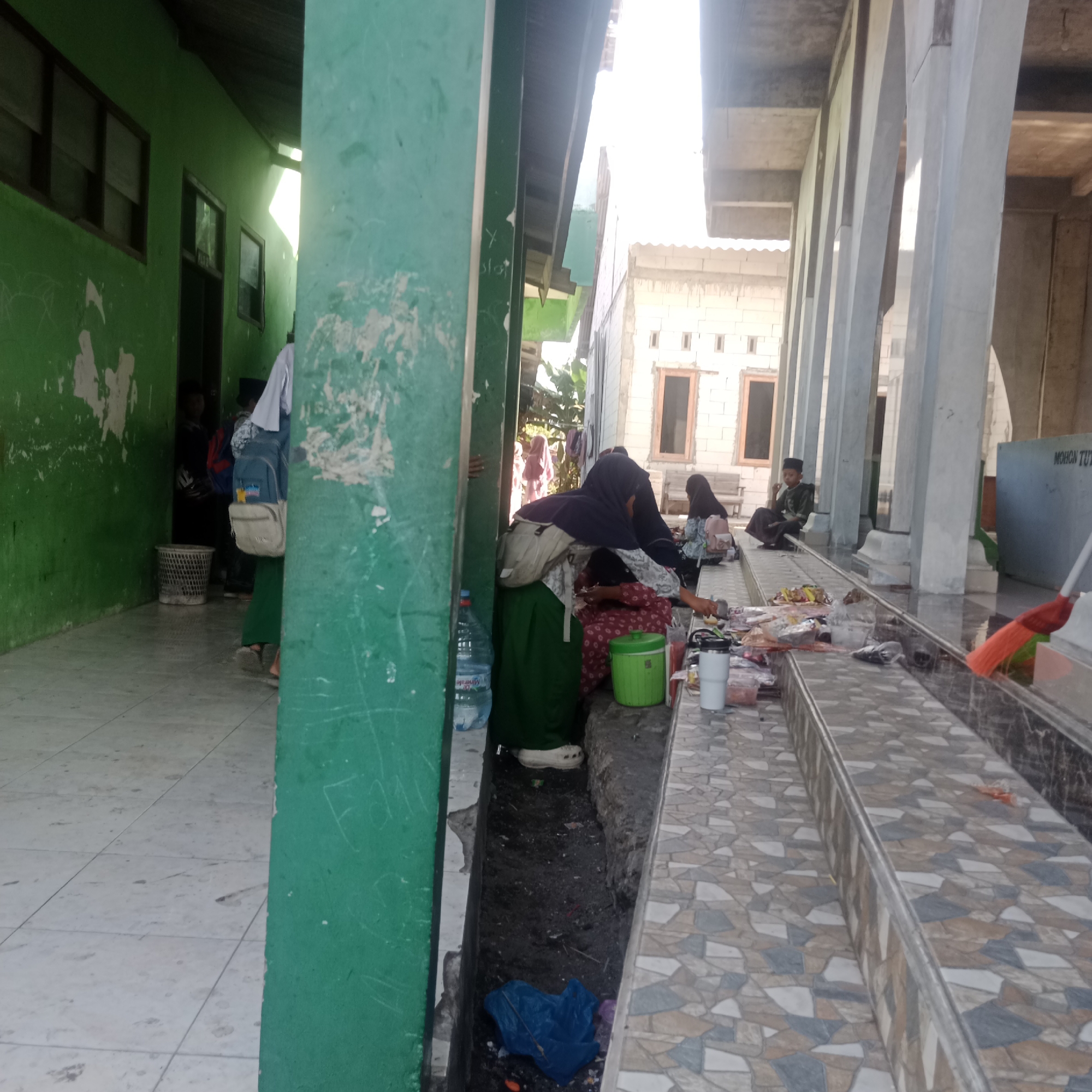 In Tegowanu Kulon, Indonesia, a group of individuals, some wearing hijabs and traditional clothing, are gathered outdoors in what appears to be a covered walkway adjacent to a building under construction. The walkway has a tiled floor and is supported by large, concrete pillars. In the foreground, various items are laid out, including plastic bags filled with trash, food containers, a water bottle, and a broom with an orange brush. A blue plastic bag lies open in a drainage ditch. To the left, a green wall with peeling paint marks the edge of the walkway, with a white trash can visible in the background. Behind the pillars, the construction of a white brick building with dark-framed windows is evident. Sunlight streams in from the right, creating bright highlights and shadows.