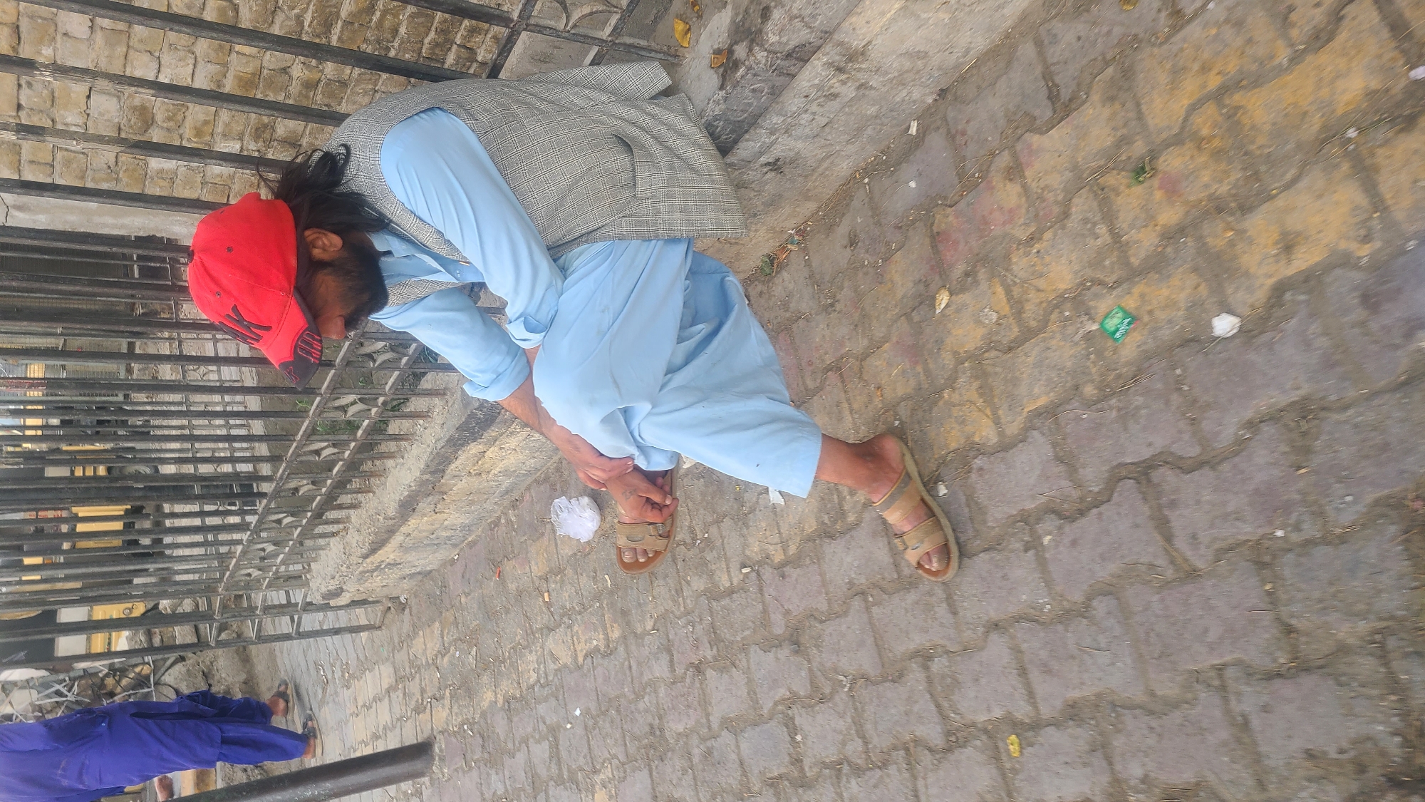 A man with a beard and long dark hair wearing a red baseball cap, a light blue shirt, a grey checkered vest, and light blue pants sits on a concrete ledge next to a metal fence. He is wearing light brown sandals. The man is looking down and to his left. To the left of the man, another person wearing a bright blue salwar kameez walks away from the camera. The ground is paved with interlocking bricks. There is litter on the ground, including a crumpled white piece of paper and what appears to be a green packet. In the background, a yellow vehicle and a black metal fence are visible. The overall scene suggests a public street or pathway in Quetta City Tehsil, Pakistan.