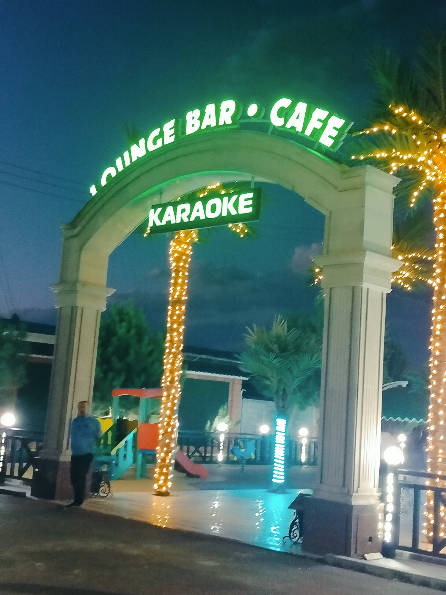 The image depicts the entrance to a "LOUNGE BAR CAFE KARAOKE" establishment at night. A man, wearing a blue shirt and dark pants, stands to the left of a large, ornate archway. The archway is illuminated with green neon lights that spell out the establishment's name. Below the main sign, a separate green neon sign reads "KARAOKE." The scene is decorated with numerous small yellow lights wrapped around palm trees, creating a festive atmosphere. In the background, a children's playground structure is visible. The ground appears wet and reflective, possibly due to recent rain or cleaning. The overall impression is of an outdoor entertainment venue in a somewhat tropical or warm climate, given the palm trees. The location is identified as Aksukent, Kazakhstan.