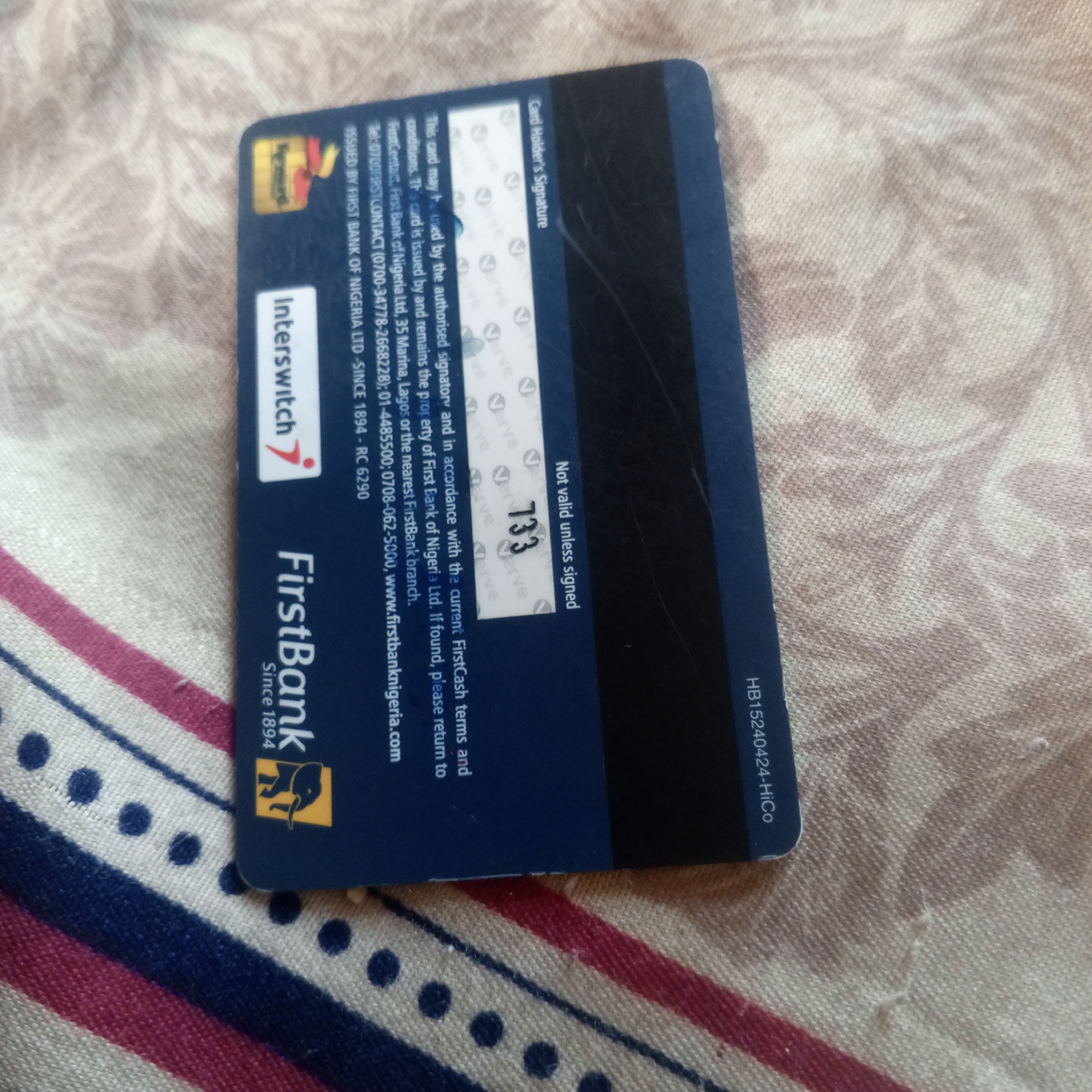 The image shows a close-up, top-down view of the back of a First Bank of Nigeria debit card. The card is dark blue with some gold and white text and logos. The text on the card includes "FirstBank Since 1894", the Interswitch logo, and a lengthy disclaimer about the card being the property of First Bank of Nigeria Ltd. and instructions for its return if found. There is a signature strip on the card with the number "733" visible, and the text "Not valid unless signed". The card is lying on a patterned fabric surface with a beige and light brown floral design. The lighting suggests an indoor setting. No people are visible in the image. The location is Unknown, Unknown.