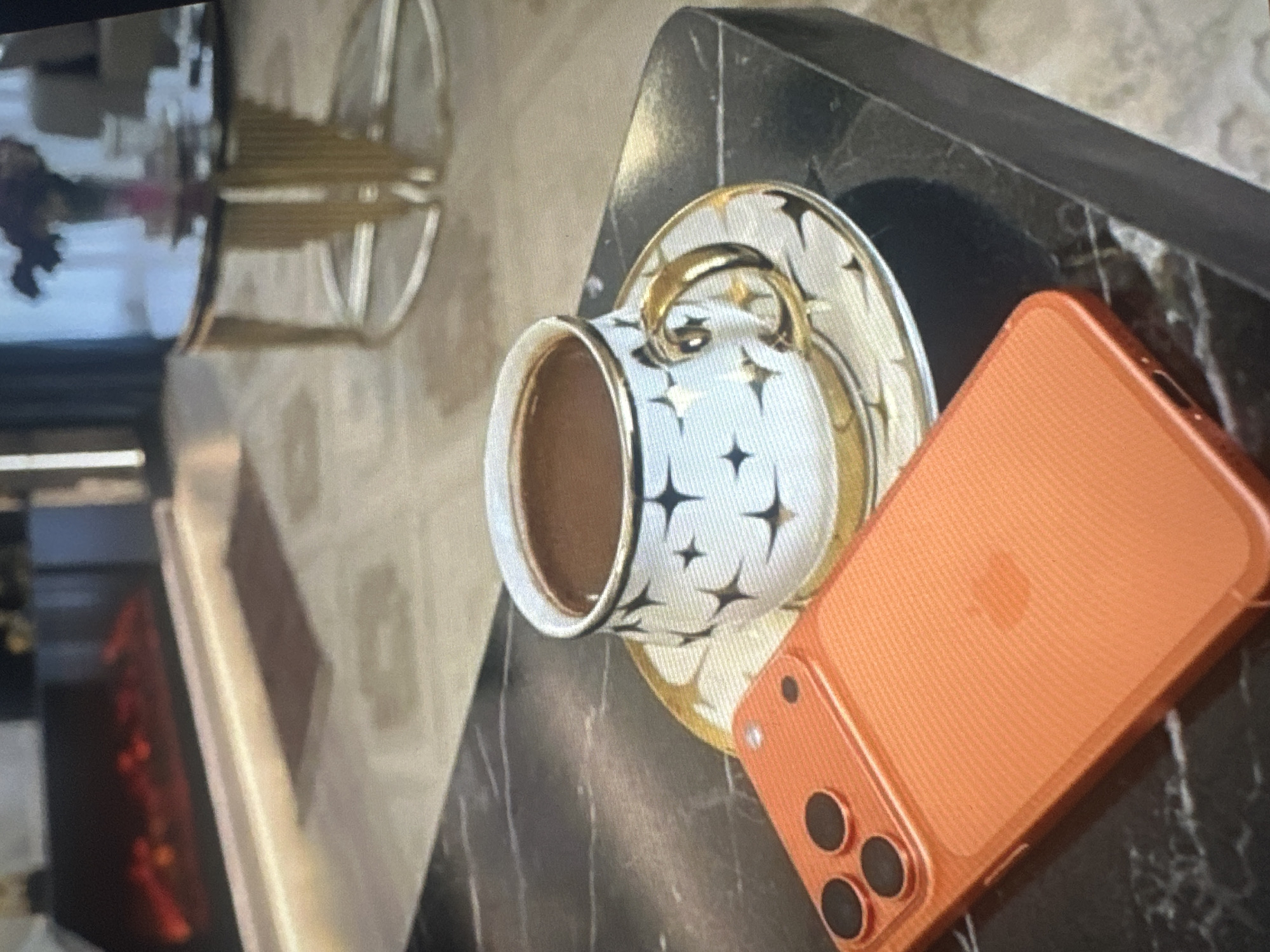 A close-up shot focuses on a dark marble table surface, upon which rests an orange smartphone and a demitasse cup filled with dark liquid, likely coffee, situated on a matching saucer. The cup and saucer feature a white background adorned with black starburst patterns and gold accents. In the background, a blurred view reveals a contemporary living space with an electric fireplace emitting a red glow, a round glass coffee table with gold detailing, and soft lighting. The scene is indoors and devoid of any discernible city or location context, including Tallinn, Estonia.