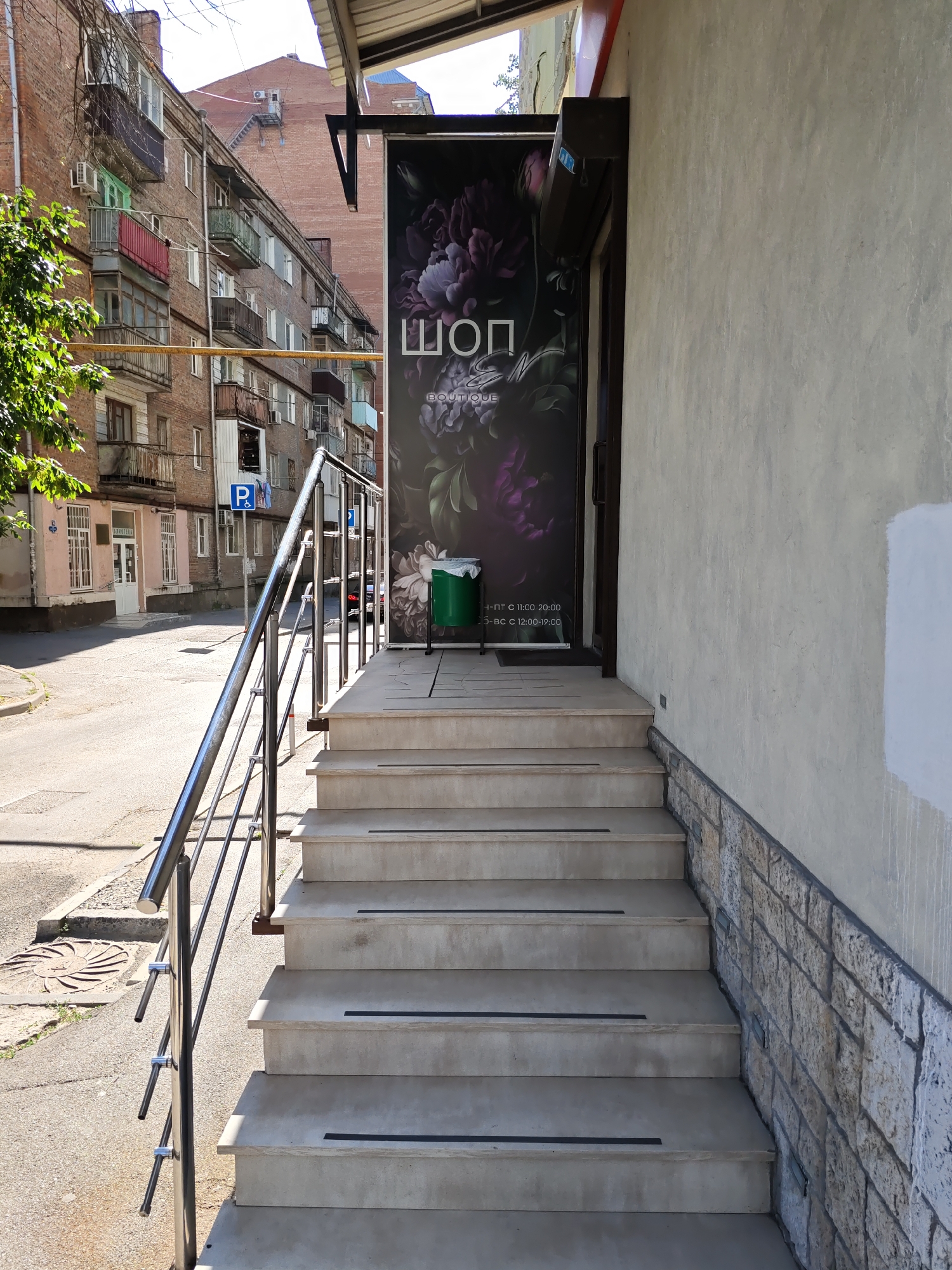 The image shows an outdoor view of the entrance to a shop called "ШОП" (SHOP) in Rostov-on-Don, Russia. The shop has a large dark-colored sign with floral imagery and text, possibly indicating business hours. A set of concrete stairs with a metal handrail leads up to the shop entrance. To the left of the stairs, a street with parked cars and apartment buildings can be seen. A green trash can is placed near the shop entrance. The architecture of the buildings suggests an urban setting. The lighting indicates it is daytime, likely with sunny conditions.