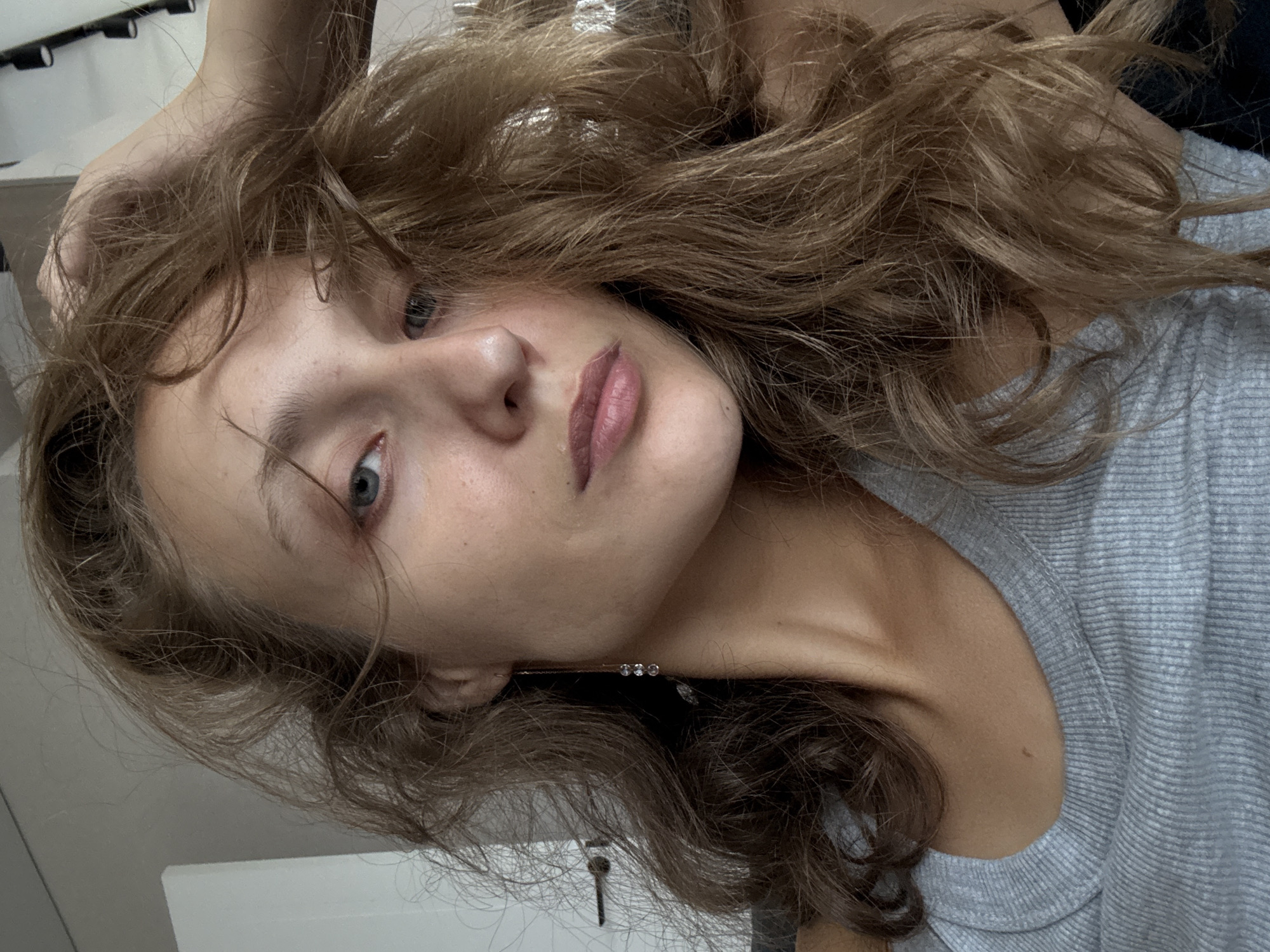 The image is a close-up, selfie-style portrait of a young woman. She has long, wavy light brown hair that is partially obscuring her face. Her left hand is raised and resting on her head, further displacing her hair. She is wearing a grey ribbed tank top. A long, delicate earring with three small clear stones is visible dangling from her left ear. The background appears to be indoors, with a white wall on the left and what looks like track lighting on the ceiling in the upper right corner. There is a faint, blurry object on the wall to the left of the woman. The overall lighting suggests a natural light source from the right. The image is devoid of any elements that would specifically place it in the city of Подшивалово, Russia, or any other specific urban location.