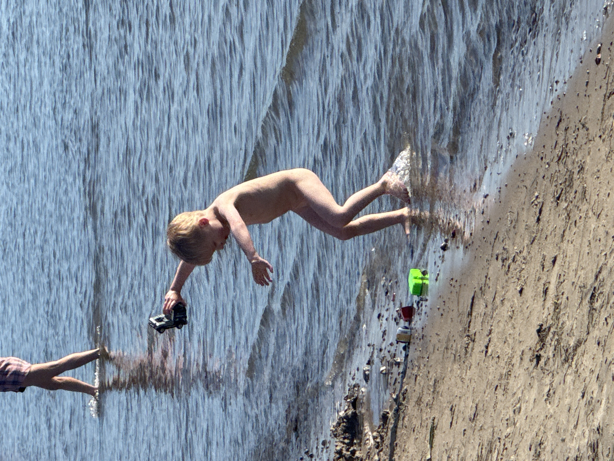 This media file depicts a playful scene on a sunny day, likely at a beach or by a body of water in Tallinn, Estonia. The main subjects are two children, one fully visible and nude, the other partially visible, wearing checkered shorts.
The nude child is captured in mid-air, appearing to be jumping or diving into the water. The child is holding a toy black car in their right hand. Their body is positioned horizontally, with legs bent and arms outstretched. The other child's legs are visible at the bottom left of the frame, reaching towards the water.
The setting is a beach with wet sand and shallow water. The water has a reflective quality, creating streaks of light and shadow that give the impression of a waterfall or cascading water. Several small toy vehicles, including a green one and a red and white one, are scattered on the wet sand near the water's edge.
The lighting suggests it is daytime, with bright sunlight casting shadows. The overall mood is one of fun and carefree childhood activity. There is no visible text in the image.