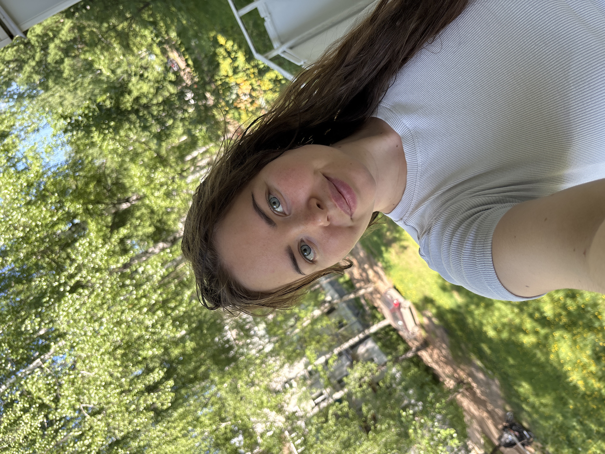 A selfie of a woman with long brown hair and blue eyes, wearing a light grey ribbed t-shirt. The background is green, featuring trees with dense foliage and patches of grass. A structure with white railings is partially visible at the top of the frame. The setting appears to be outdoors in a park or wooded area. Without additional information, I cannot confirm if the location is specifically in Izhevsk, Russia.