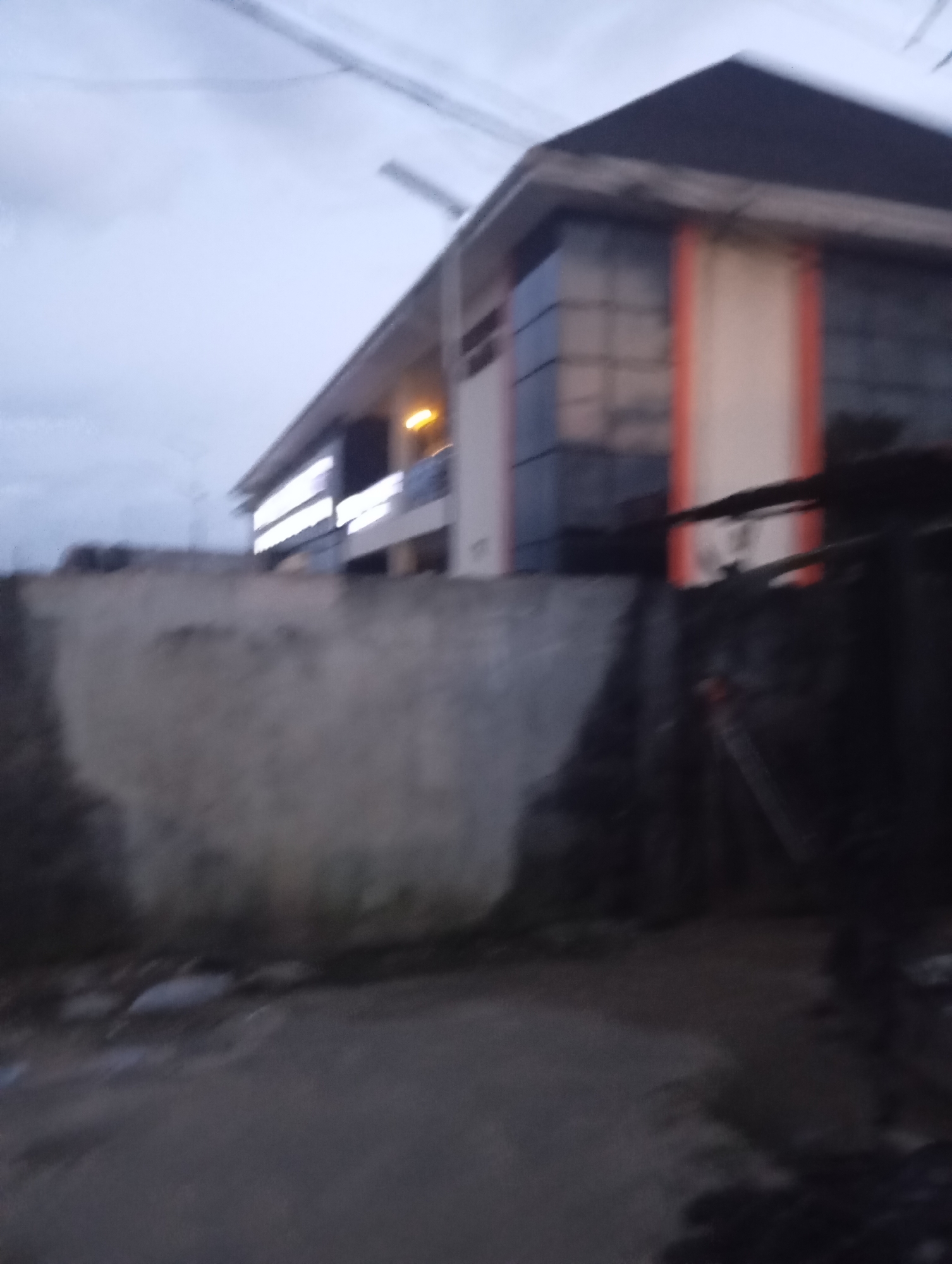 The image depicts a building with a dark roof and a facade featuring reflective glass panels interspersed with orange and white sections. Illuminated signs are visible on the lower part of the building, suggesting it may be a commercial establishment. In the foreground, a rough, weathered concrete wall runs horizontally across the frame. The ground in front of the wall appears to be a rough, unpaved surface, with some debris scattered around. The sky is overcast and the lighting suggests it is either dawn or dusk. The overall impression is of an urban setting in Port Harcourt, Nigeria, likely a street scene outside a business or office.