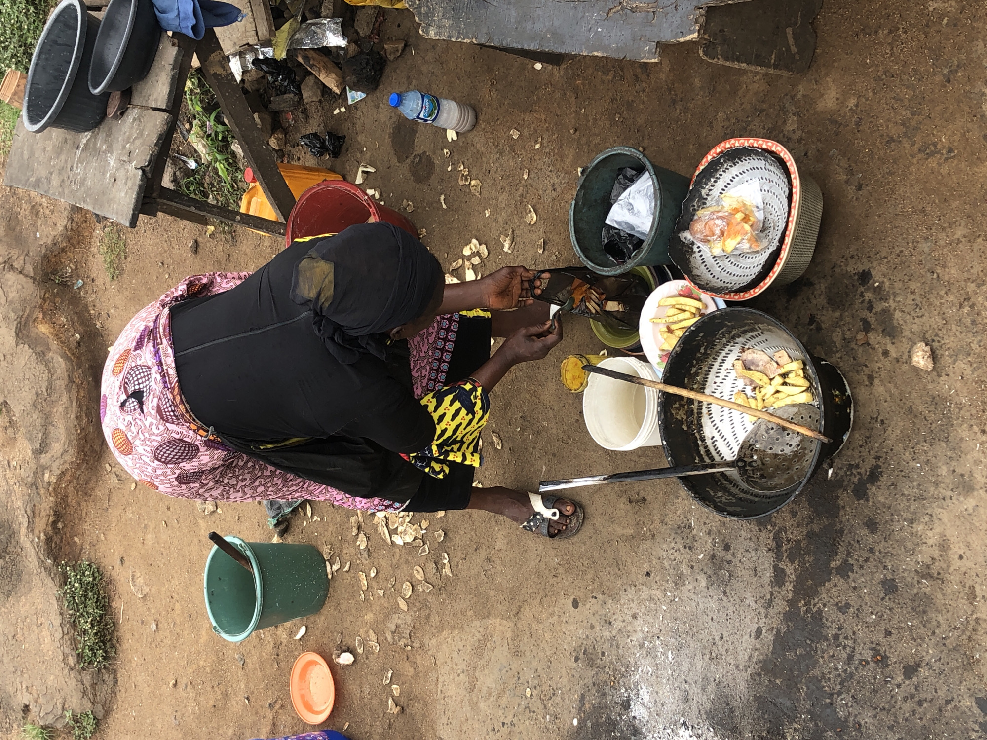 An overhead shot captures a woman in Kabong, Nigeria, tending to food preparation outdoors. She wears a black headscarf and a pink and white patterned dress over a black garment, along with a yellow and black patterned apron. The woman is bent over, holding a spoon and a black plastic bag containing food. In front of her are various containers, including a large black wok with a metal strainer holding fried food, a white bucket, and a green plastic bucket. To her right, there are several plastic bowls, one containing fried food. The ground is a mixture of dirt and debris, with scattered shells. In the background, a wooden table holds two black bowls, and a yellow plastic container. A partially filled bottle of water rests on the ground. The scene depicts a busy outdoor food stall environment.