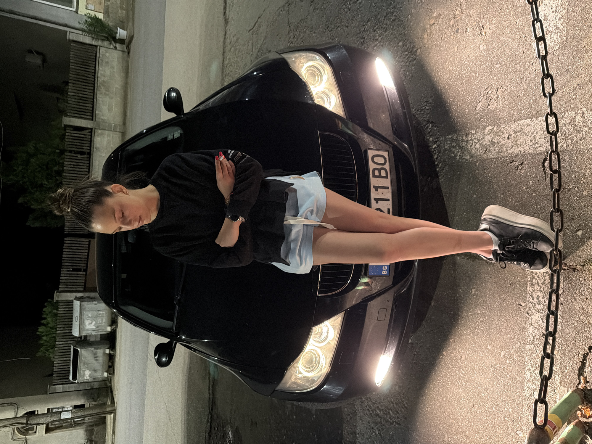 A young woman is sitting on the hood of a black BMW car. She wears a black sweatshirt, light blue shorts, and black sneakers. She has her arms crossed. The license plate of the car reads "211 BO" with the country code "BG" visible. The headlights of the car are illuminated. The scene appears to be at night, with overhead lighting on a street in Kaleytsa, Bulgaria. A chain is visible in the lower right corner. Trash containers and a building are located behind the car and the woman.