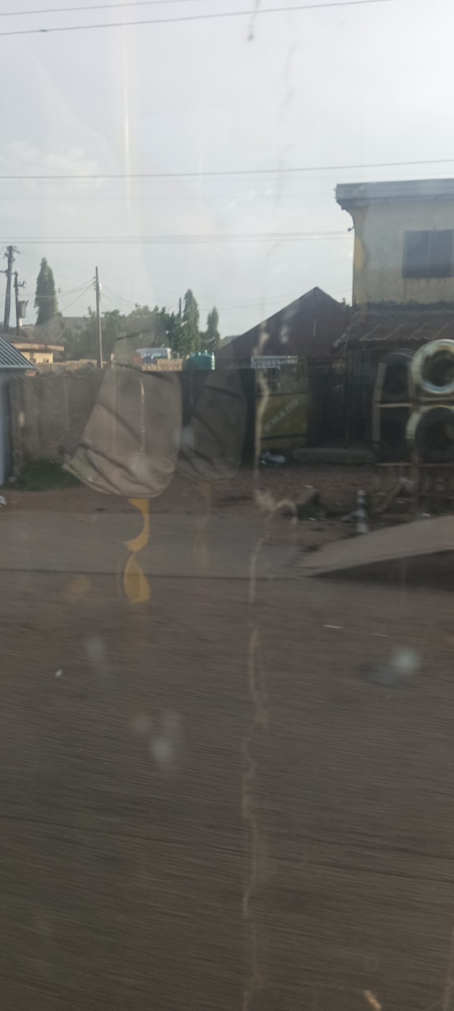 The image captures a static street view in Jos, Nigeria, taken through a reflective and somewhat smudged window, possibly from a moving vehicle. The scene depicts a mixed-use area with residential and commercial structures under a bright, hazy sky.
In the mid-ground, a low concrete wall runs horizontally, separating the foreground from several buildings. To the right, a structure with a dark, perhaps corrugated metal roof and a visible dark window is present. Next to it, another building features a yellow facade or sign with partially visible dark text. On the far right, a rack holds several stacked tires, indicating a tire-related business. Utility poles with power lines crisscross the upper portion of the frame, and slender trees are scattered in the background, behind the buildings. The ground appears uneven, a mix of dirt and rough surfaces.
A key detail is the visible text "MICHELIN TYRE" (or "TIRE") on the yellow surface of one of the buildings, directly linking to the stacked tires and suggesting a tire sales or repair shop.
No specific activity or people are clearly visible. The time of day appears to be late morning or early afternoon, given the bright, somewhat diffuse lighting and lack of harsh shadows. The sky is largely overcast or hazy. The reflections and smudges on the window obscure the immediate foreground, adding a slightly dreamlike or distant quality to the view. The overall impression is that of an everyday urban or semi-urban streetscape.