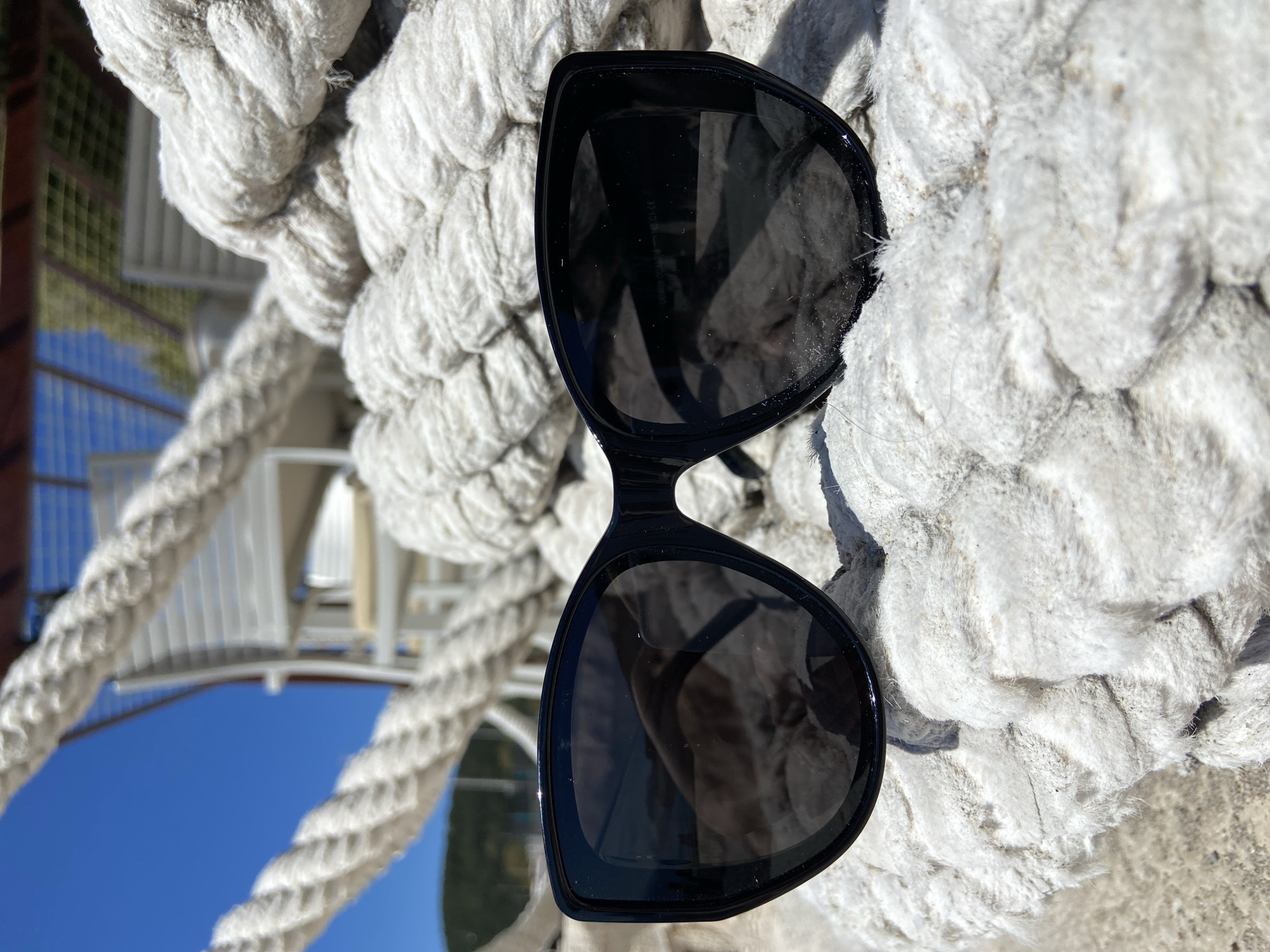 A pair of black sunglasses rests on a thick, white rope. The rope appears to be part of a larger nautical structure, possibly on a boat or dock. A partial view of white metal structures and a bright blue sky can be seen reflected in the lenses of the sunglasses. The location is likely Prijevor, Montenegro, suggested by the image content, suggesting a maritime or coastal environment.