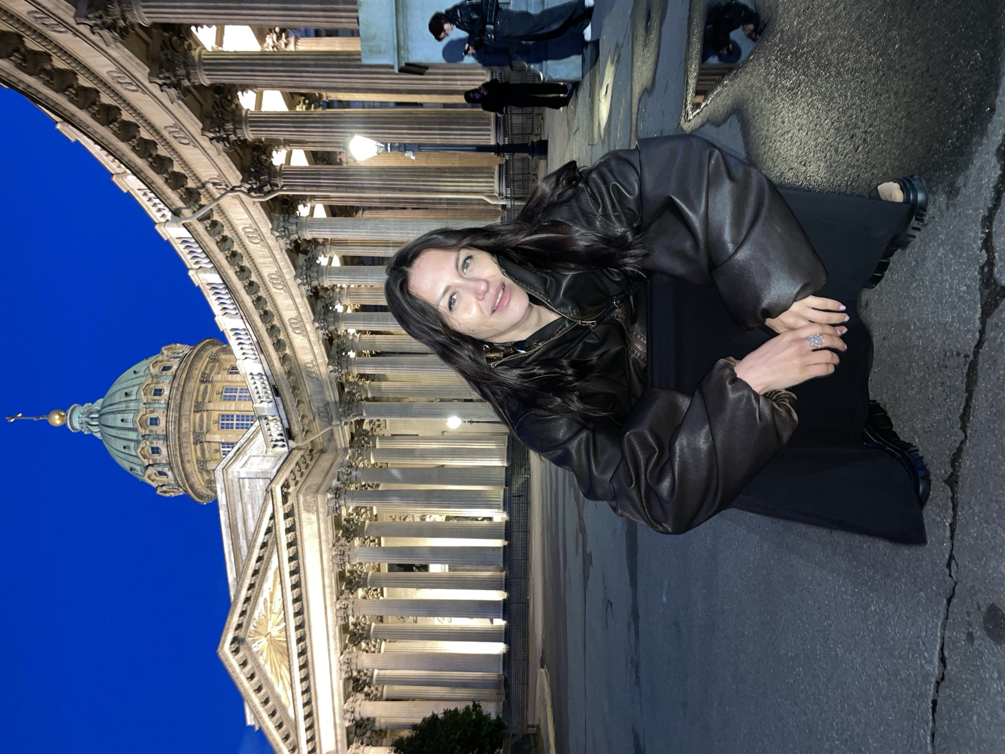 The image shows a woman crouching in front of Kazan Cathedral in Saint Petersburg, Russia. She is wearing a dark leather jacket and black pants. The cathedral, a prominent landmark in the city, is illuminated by artificial lights against the night sky. The woman is looking towards the camera with a slight smile. In the background, several other people are visible, and there are streetlights and architectural features of the cathedral. The ground in front of the woman appears to be wet, possibly from recent rain, reflecting the surrounding lights and structures.