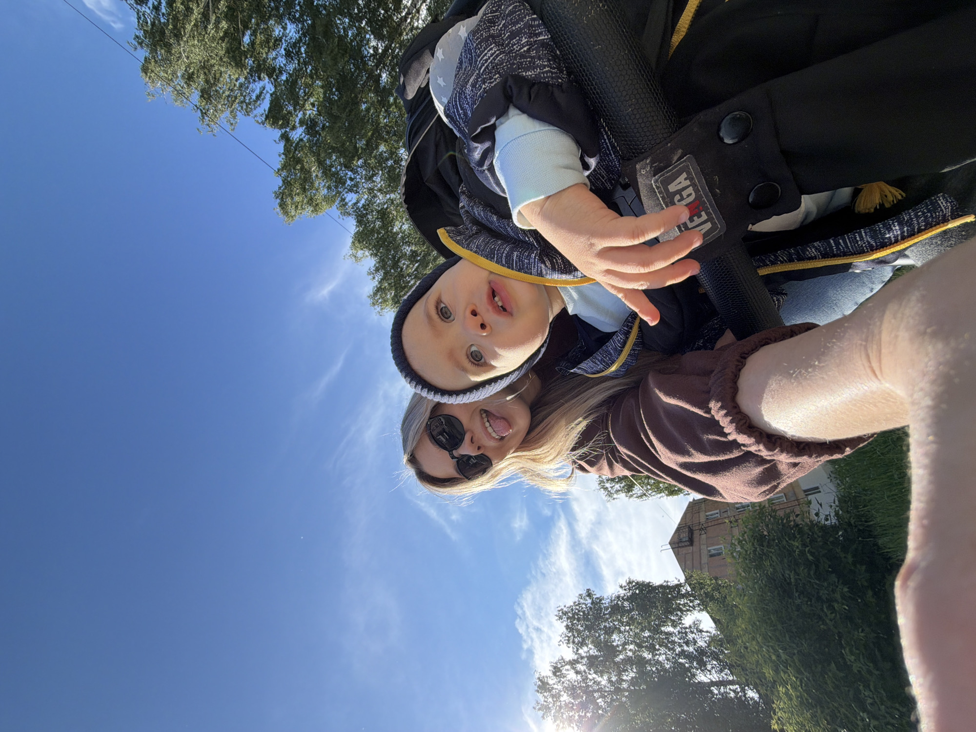 A selfie of a fair-skinned woman and a fair-skinned child against a background of a blue sky with light clouds and greenery. The woman, wearing sunglasses, is sticking out her tongue. The child, wearing a hat, looks directly at the camera with a surprised expression. The woman is wearing a brown, long-sleeved shirt, and the child is bundled in a black, blue, and white jacket. Part of what appears to be a black baby carrier with "VEGA" is visible. In the background, a building can be seen, along with trees. It is in the city of Chaikovsky, Russia.