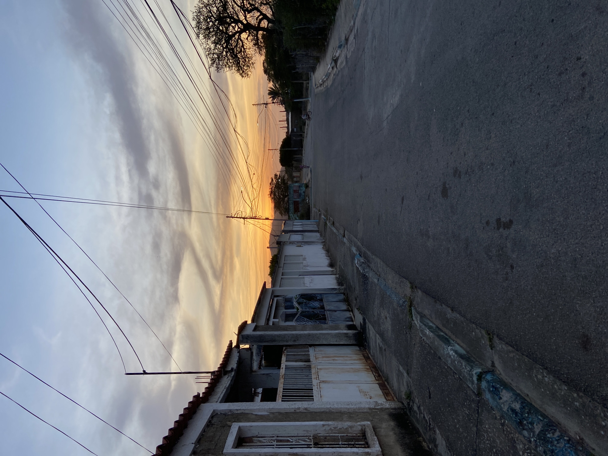 The photograph shows a street scene in Juan Griego, Venezuela, taken at sunset. The road stretches into the distance, flanked on one side by low buildings with tiled roofs and on the other by a curb with some sort of painted surface. Electrical wires crisscross above the street. The sky is filled with clouds tinted orange and yellow by the setting sun. In the background, silhouetted trees and telephone poles can be seen. A person on a bicycle and another individual walking are also visible in the distance.
