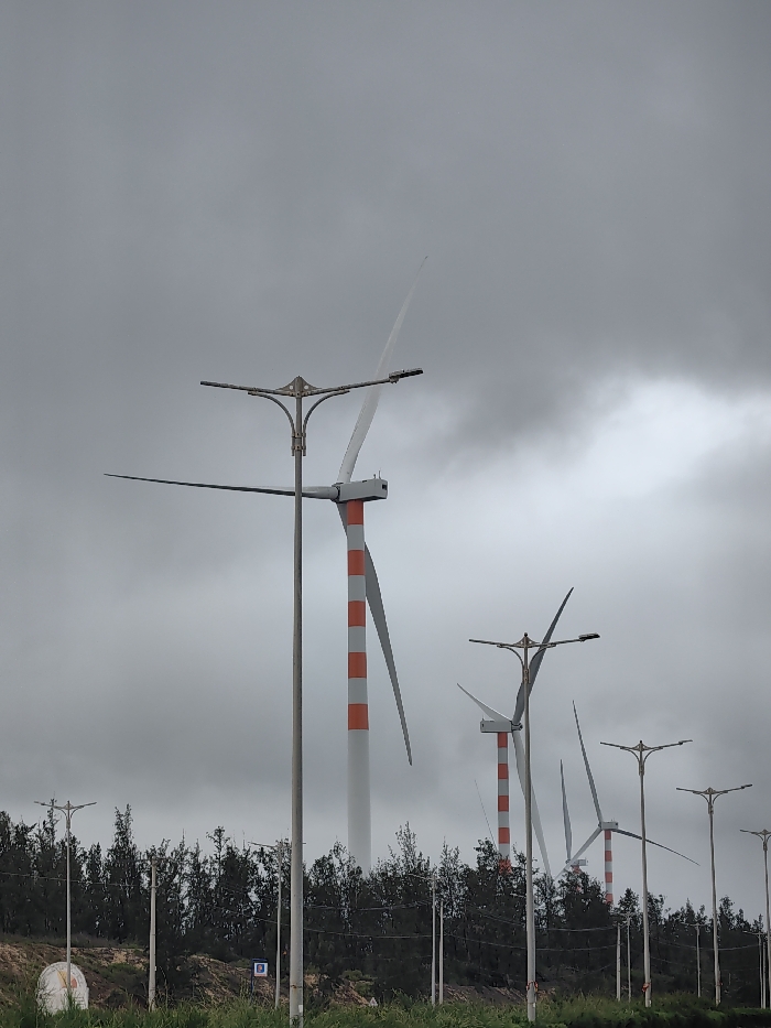 The image displays a wind farm located in East Quy Nhon Ward, Vietnam, under an overcast, grey sky. Multiple utility-scale wind turbines are visible, characterized by tall, cylindrical towers with alternating orange-red and light grey horizontal stripes, supporting three white or light grey blades. The most prominent turbine, positioned in the mid-left, has two blades partially within the frame, with one extending upwards and the other horizontally to the left. Other turbines are visible in the background, appearing smaller and partially obscured.
In the foreground and mid-ground, several metallic grey streetlights are present, each featuring a central pole topped with four radiating arms that support horizontal light fixtures. One such streetlight stands directly in front of the largest wind turbine, partially overlapping its structure.
The lower portion of the scene consists of a strip of green grass, followed by an unpaved earthen embankment, and then a dense horizontal line of dark green, slender trees or bushes. A small, partially visible white spherical or dome-shaped object rests on the ground at the far left, and a small blue sign with white symbols is present near the center-left, at ground level. No individuals or animate subjects are depicted, nor are any specific actions or interactions taking place.