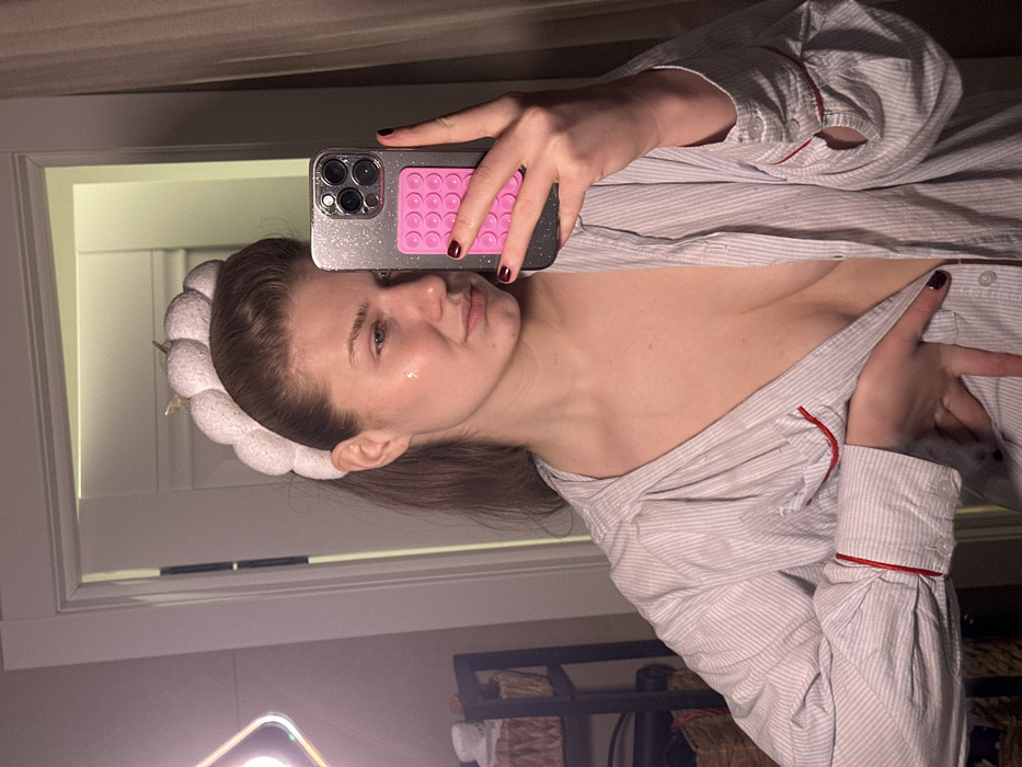 The image displays a mirror selfie of a female individual, observed indoors. The described location is the city of Yanga-Yul, Russia.
The subject, a female with light brown hair, wears a white, textured headband with a subtle golden accent. A reflective sheen is present on her left cheek. She is dressed in a light-colored, vertically striped button-down shirt, unbuttoned to expose her upper chest. The shirt features long sleeves with red piping visible on the cuffs. Her fingernails are painted with dark maroon polish. She holds a smartphone, identifiable by its multi-lens camera array and a glitter-finish case, in her right hand. A pink, multi-suction cup accessory is affixed to the phone's back. Her left hand is positioned over her midsection, partially obscuring the shirt's buttons. Her expression is neutral as she looks towards the mirror.
The reflected environment includes a white door frame behind the subject. To the far left, a square-shaped, illuminated LED panel is partially visible. Adjacent to the light, a dark-colored, multi-tiered shelving unit or basket stand is partially visible, containing woven baskets. A light-colored curtain or wall is present on the far right.