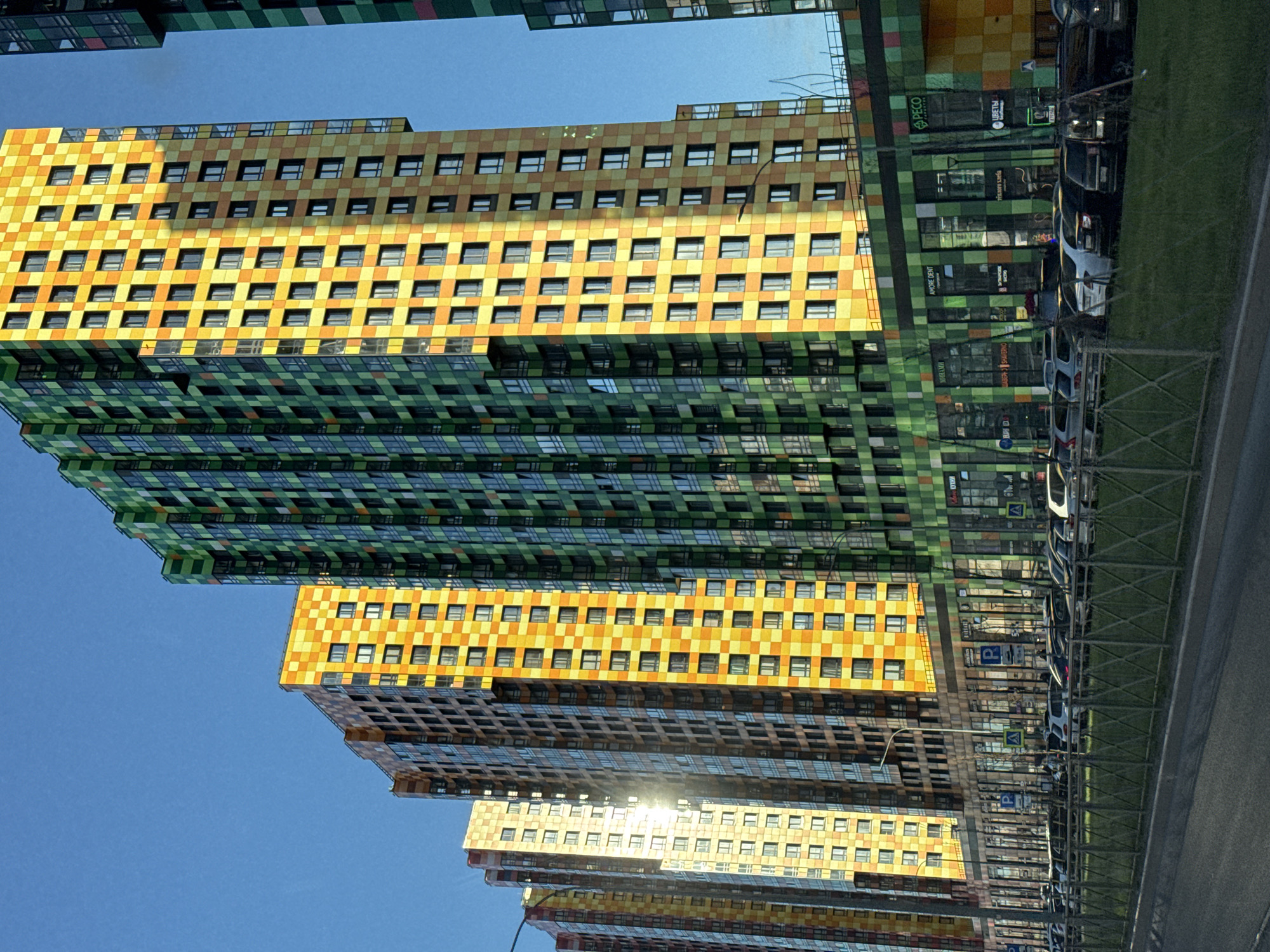 The image shows an upward view of a series of colorful, modern apartment buildings in Saint Petersburg, Russia. The buildings are predominantly yellow and green, with a checkerboard pattern on their facades. Each floor of the buildings features rows of rectangular windows. The sky is clear and blue, and the sun reflects off some of the buildings. A row of parked cars runs alongside the buildings, separated by a metal fence and grassy area. Signs with the word "PECO" are visible on the buildings, suggesting commercial spaces.