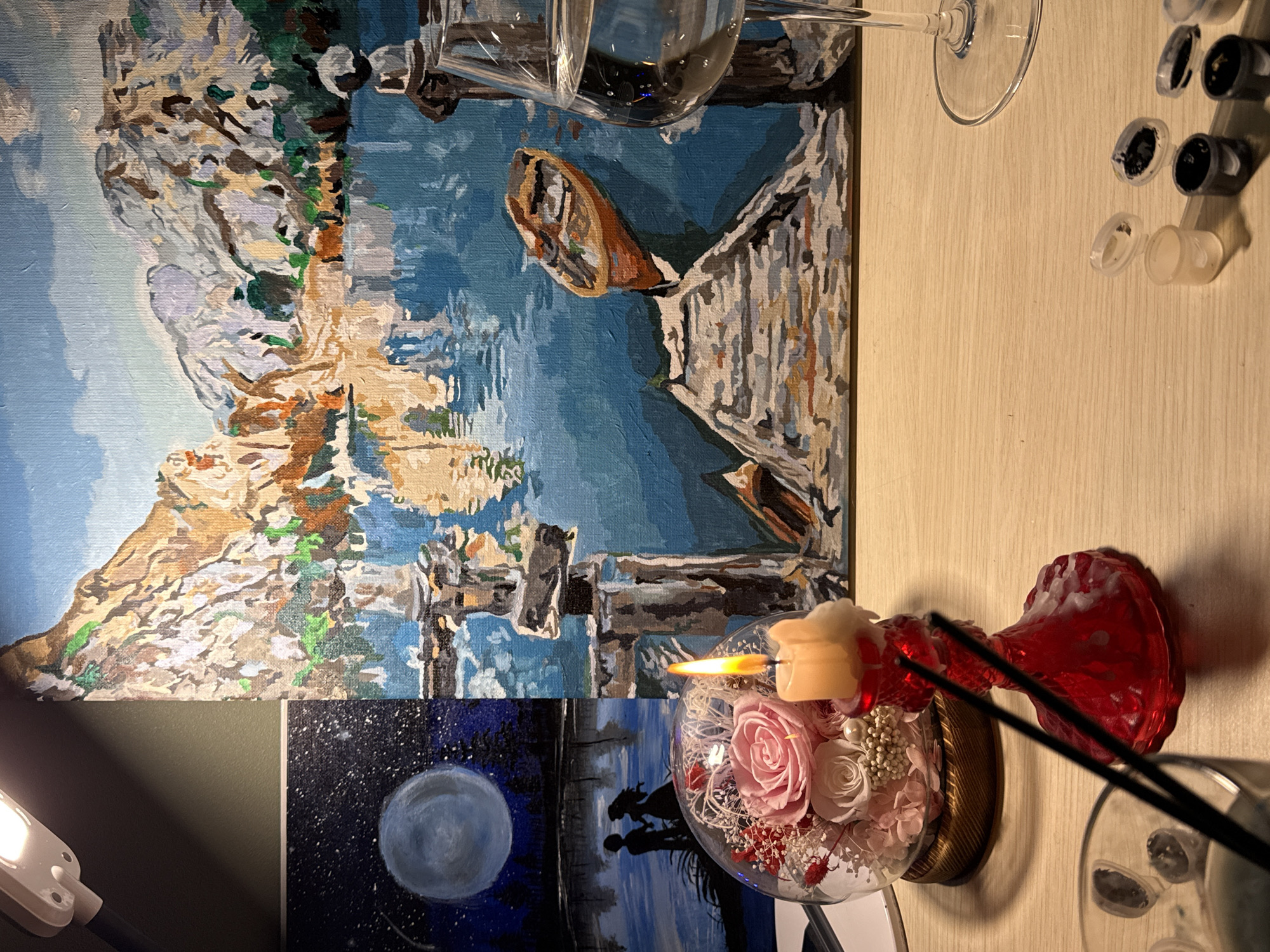 A close-up, high-angle shot shows a table with art supplies and a lit candle. The main focus is a paint-by-numbers canvas depicting a lake with mountains and a small boat. To the left, another canvas shows a moonlit night scene with two silhouetted figures. A glass dome with preserved flowers sits next to a red candle holder with a lit candle. Several small paint pots are scattered on the table. A glass of clear liquid is positioned to the right of the main canvas. The lighting suggests an indoor setting, possibly a home or studio. The location of Cheboksary, Russia, is not discernable from the image.