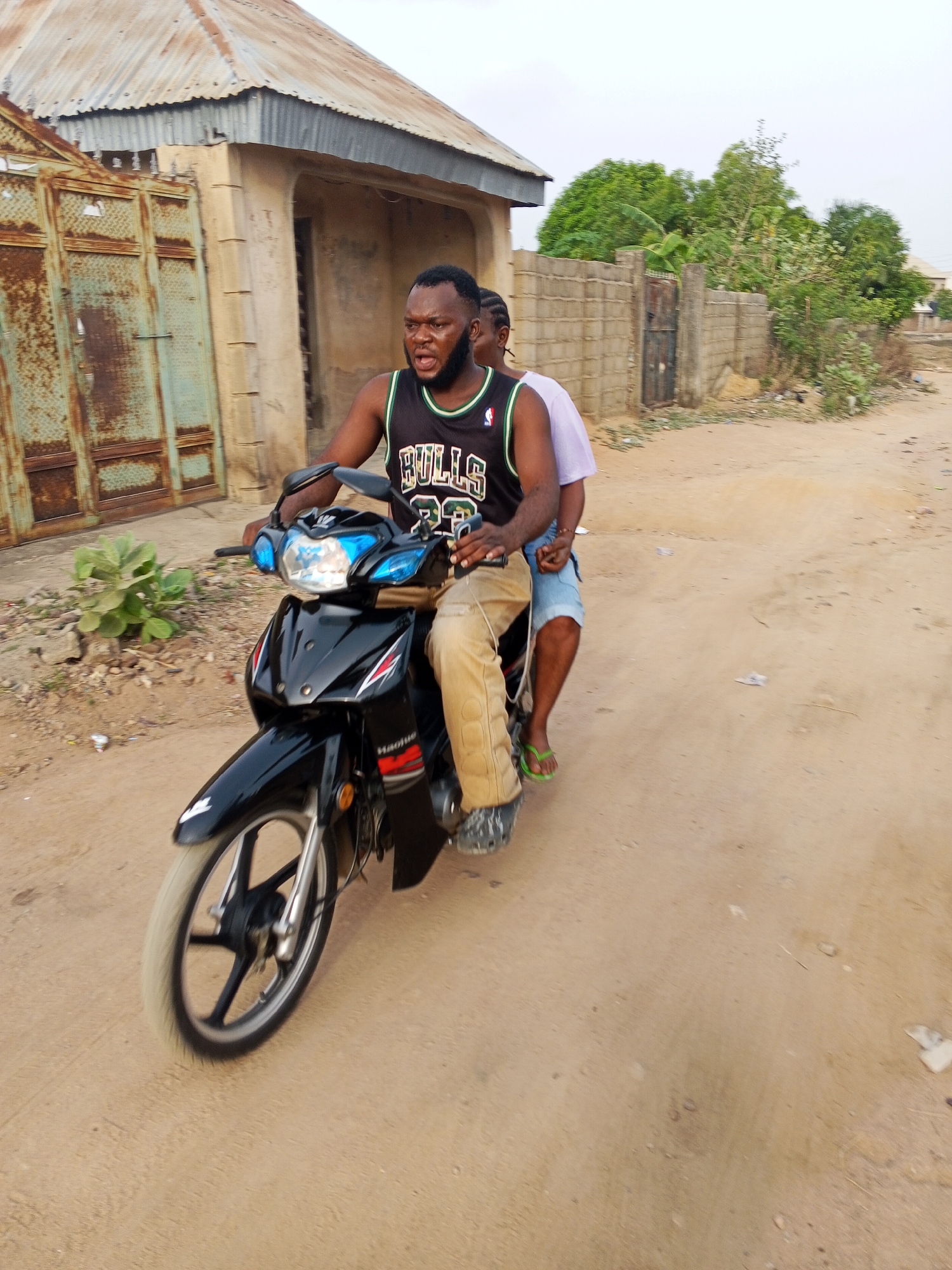 Two people are riding a black motorcycle down a dirt road in Adonkolo, Nigeria. The rider is a dark-skinned man with a beard, wearing a Chicago Bulls jersey and khaki pants. The passenger is a dark-skinned person in a light purple shirt and blue shorts. The road is unpaved, and there is a building with a rusty metal gate on the left side of the frame. Trees and a wall are visible in the background.