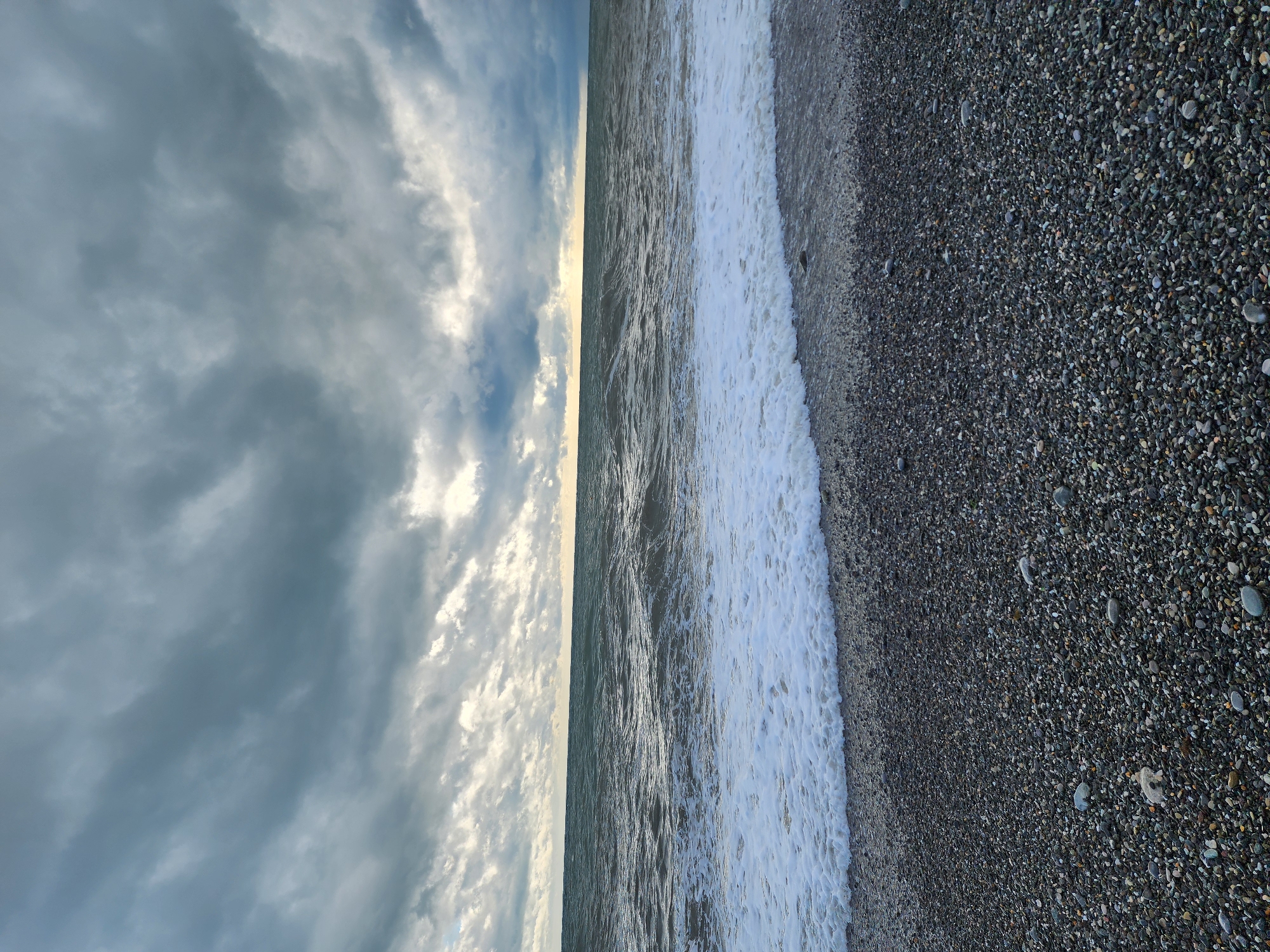 This is a photograph of a coastline in Sochi, Russia. The view is split between a cloudy sky on the left, a body of water in the middle, and a pebble beach on the right. The waves are crashing on the shore. The overall lighting suggests either dawn or dusk. There are no people visible in the scene.