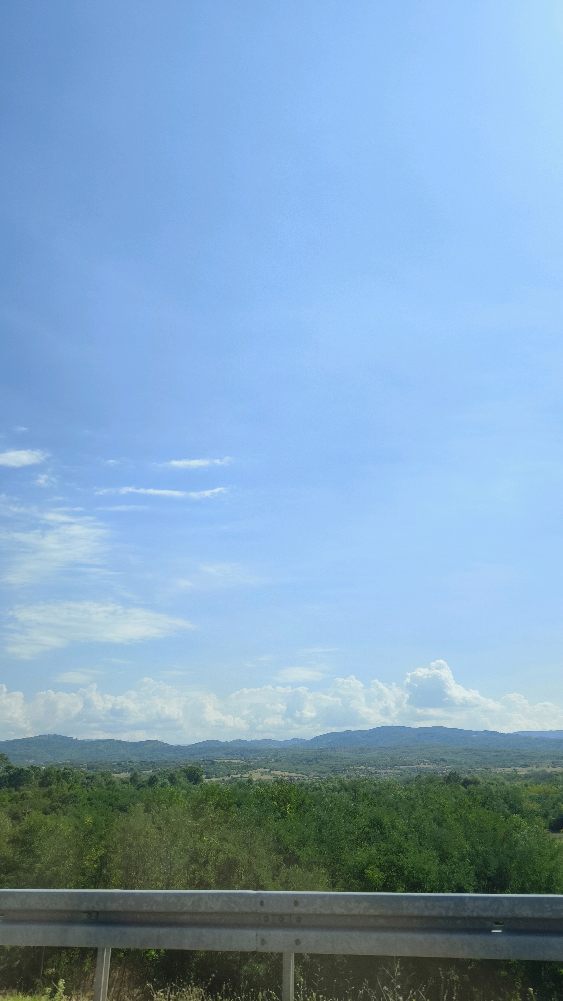 The image captures a vast, clear blue sky with scattered wispy clouds. Below the sky, a dense, green forest stretches across rolling hills and distant mountains. In the foreground, a silver metal guardrail runs horizontally across the bottom of the frame, suggesting a roadside vantage point. The scene is devoid of any people or specific structures that would pinpoint a location within Vranje, Serbia. The environment is natural and undeveloped, featuring abundant vegetation and a panoramic view of the landscape under a bright sky.