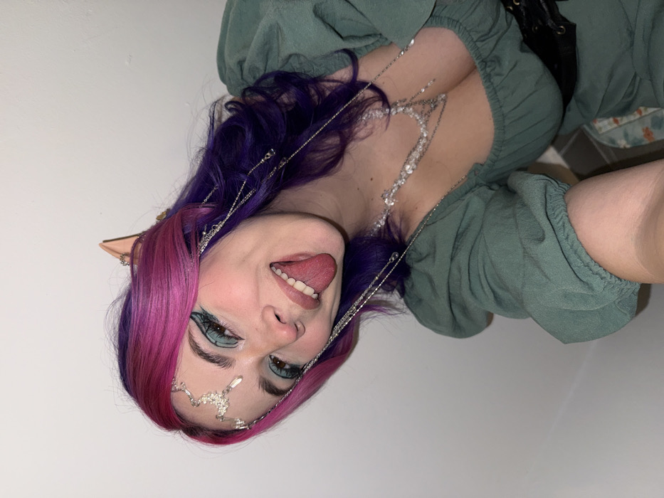 A woman is shown from the chest up. Her hair is dyed pink and purple and she is wearing elf ears. She has on green eye makeup and a decorative silver headpiece with small rhinestones. She is wearing a green peasant top and a black corset. Her tongue is sticking out and she has on a silver rhinestone necklace. The picture is rotated counterclockwise.