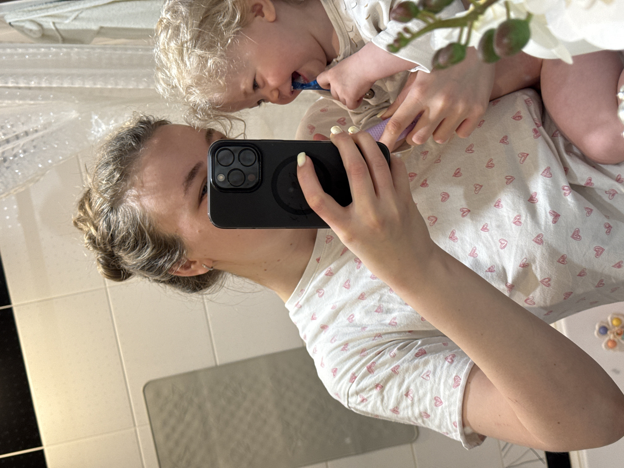 Here is a detailed description of the image:
The image captures a mother in a bathroom setting, presumably taking a selfie in a mirror. She is holding a black smartphone up to her face with one hand, seemingly capturing the reflection. In her other arm, she holds a young child who is in the process of brushing their teeth. The child's small hand is visible, holding a purple toothbrush.
The mother is wearing a white shirt with a pattern of pink hearts. Her hair is styled up and back. The bathroom features white tiles on the walls and a gray mat on the floor. A black strip runs along the base of the wall on one side.
There is also a set of artificial white flowers in the upper right. The picture appears to have been taken close to the mirror.
