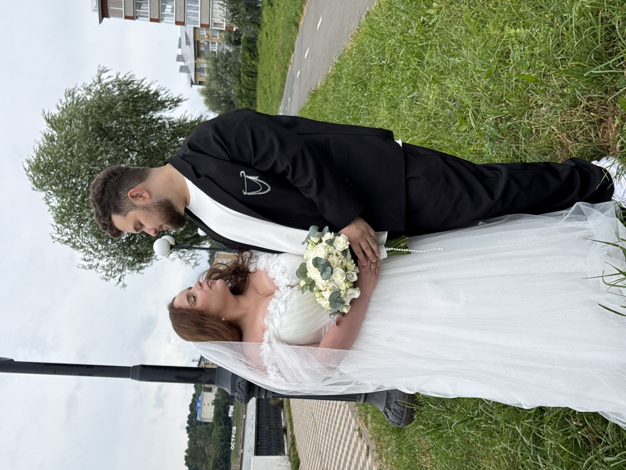 A newly married couple stands on a grassy area next to a paved walkway. The woman is wearing a white wedding dress and veil, holding a bouquet of white roses. The man is in a black suit jacket and pants, with a white t-shirt underneath. They are facing each other and appear to be looking into each other's eyes. In the background, there is a street lamp, a body of water, buildings, and trees. The location is the embankment of the Selyevsky pond in Votkinsk, Russia.