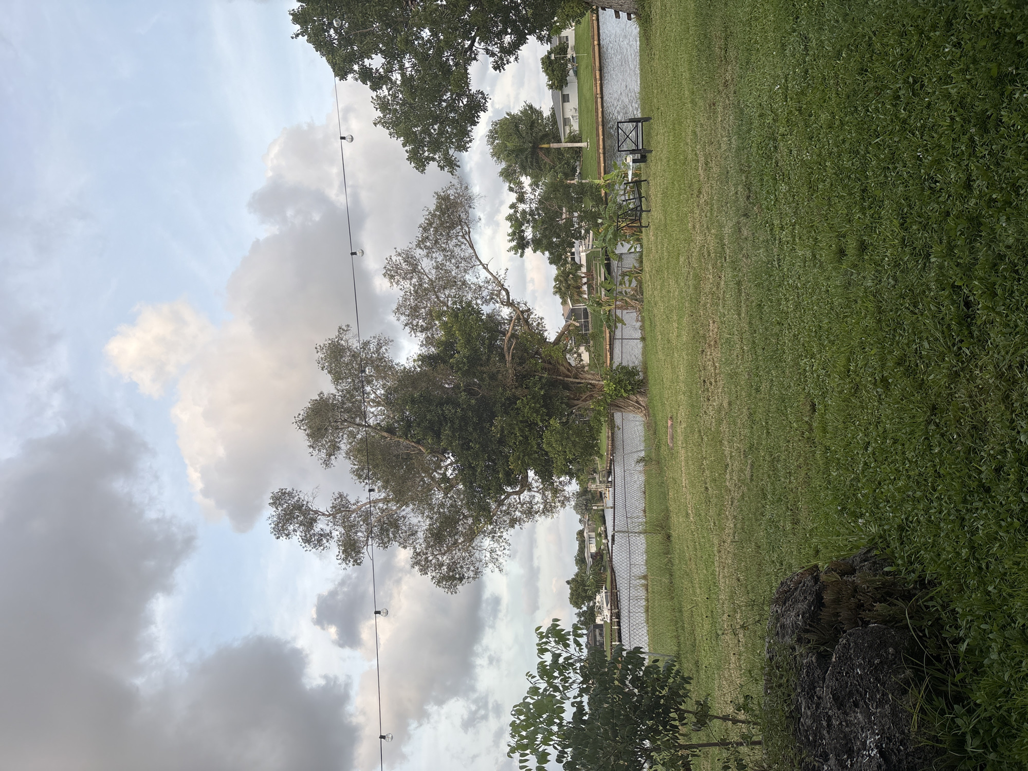 The image depicts a waterfront view in North Port, Florida. In the foreground, a grassy lawn slopes down towards a canal. To the left, a large, dark rock formation is visible, partially covered in vegetation. Two black metal chairs and a small table with a white top are set up on the grass near the canal, suggesting an outdoor seating area. A chain-link fence runs along the edge of the property, separating it from the canal.

The canal itself is calm and reflects the sky, with several boats moored on the opposite bank. Residential homes and palm trees are visible in the background across the water. The sky is partly cloudy with large, dramatic cloud formations, indicating either early morning or late afternoon light. String lights with visible bulbs are strung across the top of the frame, adding an element of an outdoor living space. The overall scene suggests a residential area in North Port, likely a neighborhood situated along a waterway.