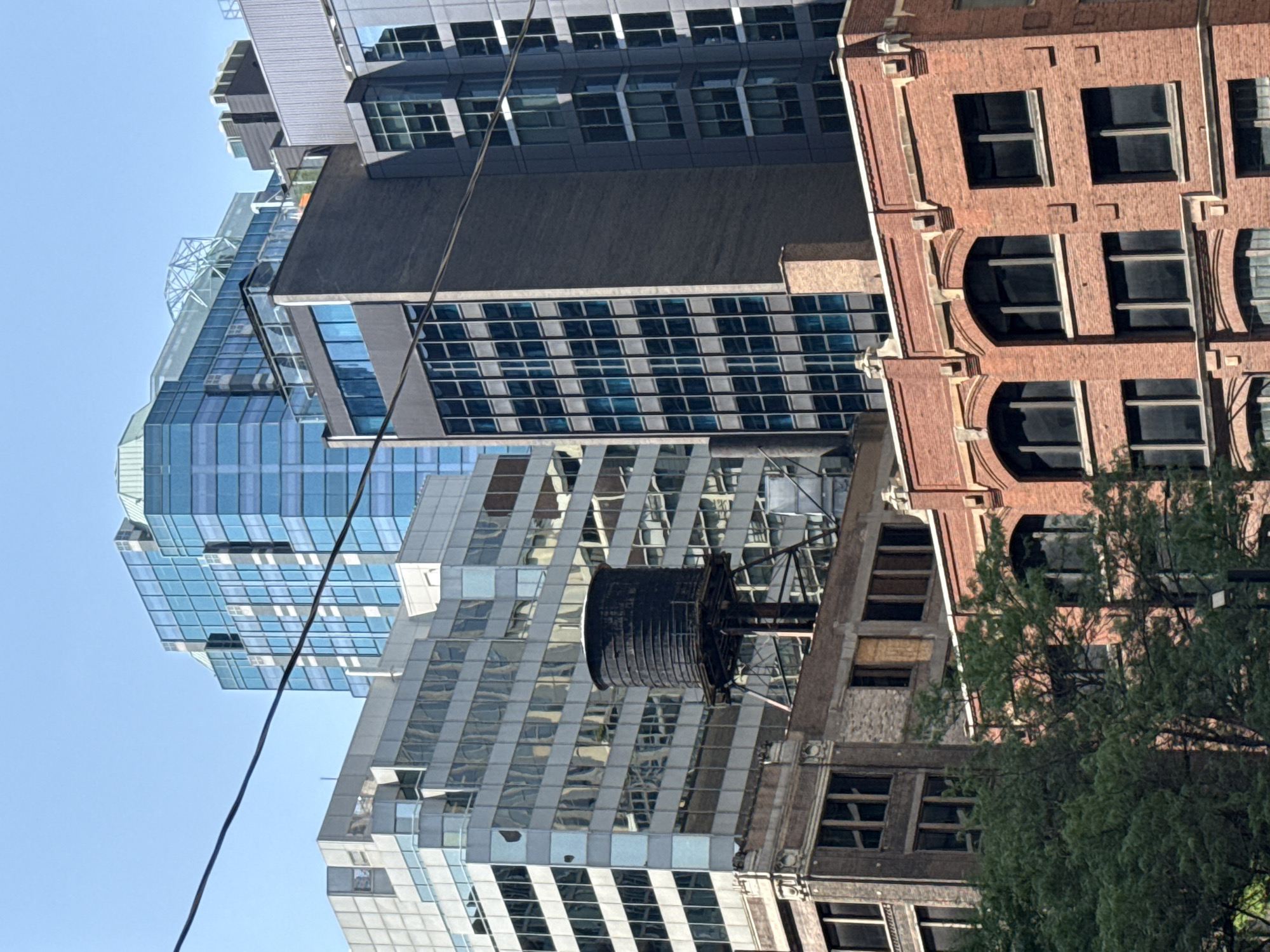 The image depicts a view of the Chicago skyline on a clear day, with a variety of buildings of different architectural styles and heights. In the foreground, a red brick building with arched windows and decorative stone elements is visible, partially obscured by lush green trees. Above and behind this, a darker brick building with a simpler facade and large windows can be seen. The middle ground is dominated by a large, modern building with a glass facade that reflects the sky and surrounding structures, and a prominent black water tower perched on a metal frame. Further in the background, a tall, blue glass skyscraper with a distinctive faceted design rises above the other buildings, its structure reaching towards the clear blue sky. Overhead, dark electrical wires stretch across the frame, adding a utilitarian element to the urban landscape. The scene suggests a dense urban environment characterized by a mix of historical and contemporary architecture.