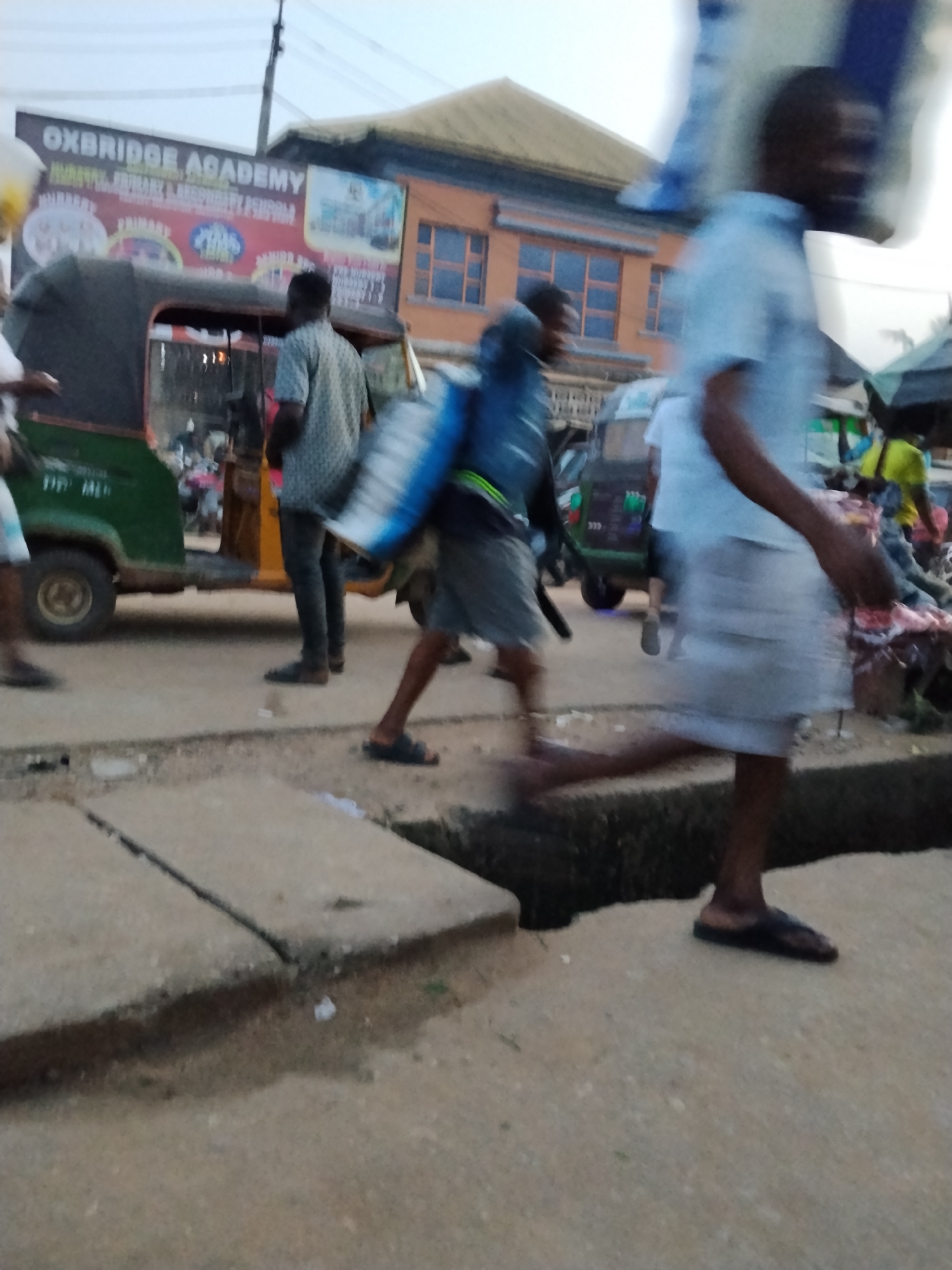 The image captures a street scene in what is likely Ura Nta Umuarandu, Nigeria, suggested by the "OXBRIDGE ACADEMY" sign in the background. The scene is composed of a partially obscured urban landscape with people in motion. In the foreground, a blurred figure is seen stepping over a concrete channel or drainage. To the left, a person wearing dark pants and a patterned shirt stands by a green and yellow three-wheeled vehicle (likely a "keke" or auto-rickshaw). Another figure is carrying a large bundle on his back. Further back, other figures and another keke can be observed, suggesting a busy market or transportation hub. The building in the background is a multi-story structure with an orange facade. The general environment seems congested with pedestrians and vehicles, possibly during daytime, given the ambient light.