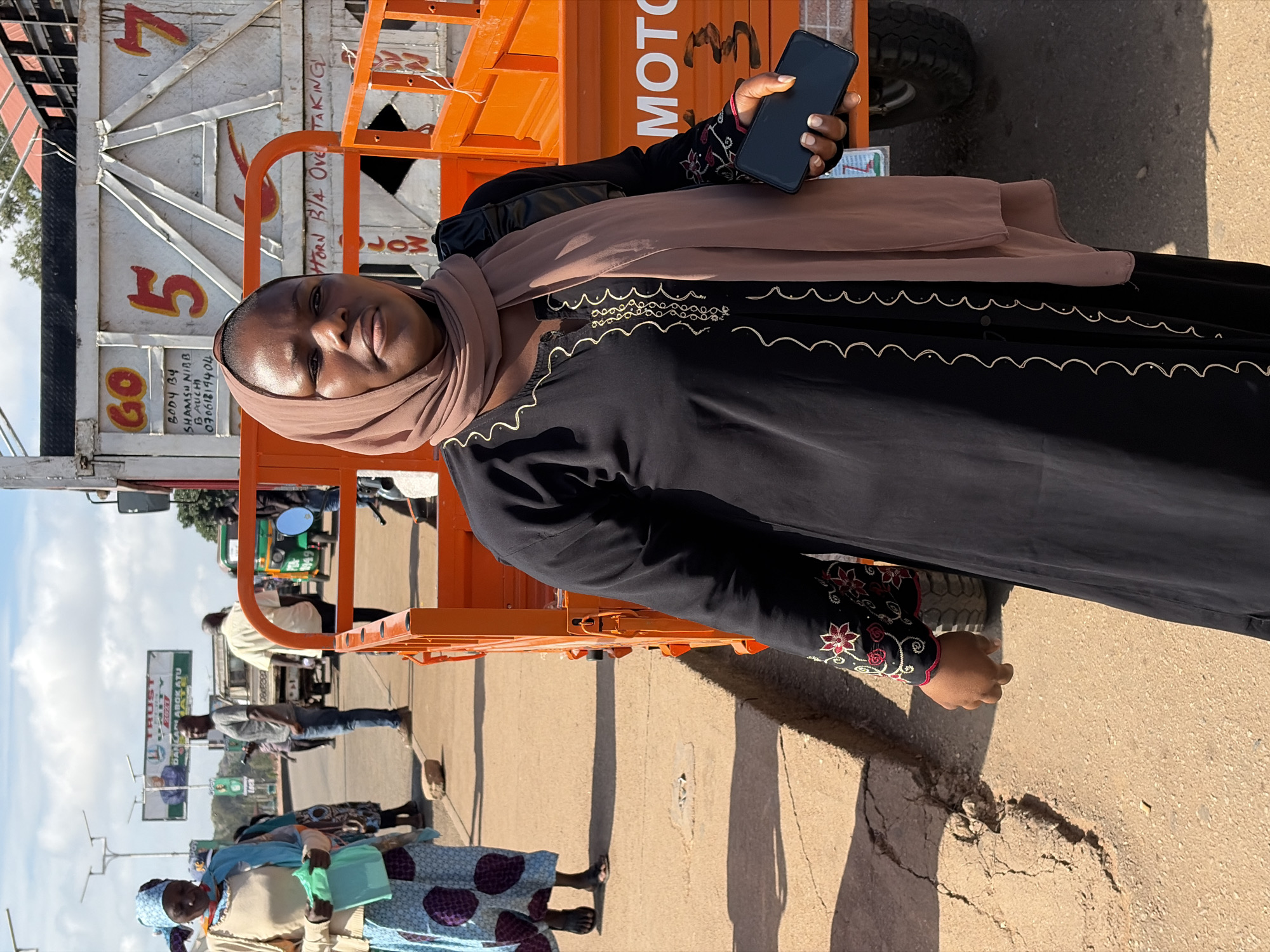 In Gura Top, Nigeria, a woman in a black abaya with gold trim and a pink headscarf stands in front of an orange flatbed truck. She holds a black smartphone in her right hand. The truck has the number "5" and "7" painted on its side. To the left of the truck, another woman in a blue and white patterned skirt and a beige cardigan holds a green bag. Several other people are visible in the background, along with a small green auto-rickshaw and a billboard in the distance. The scene takes place on a paved road under a clear, sunny sky.
