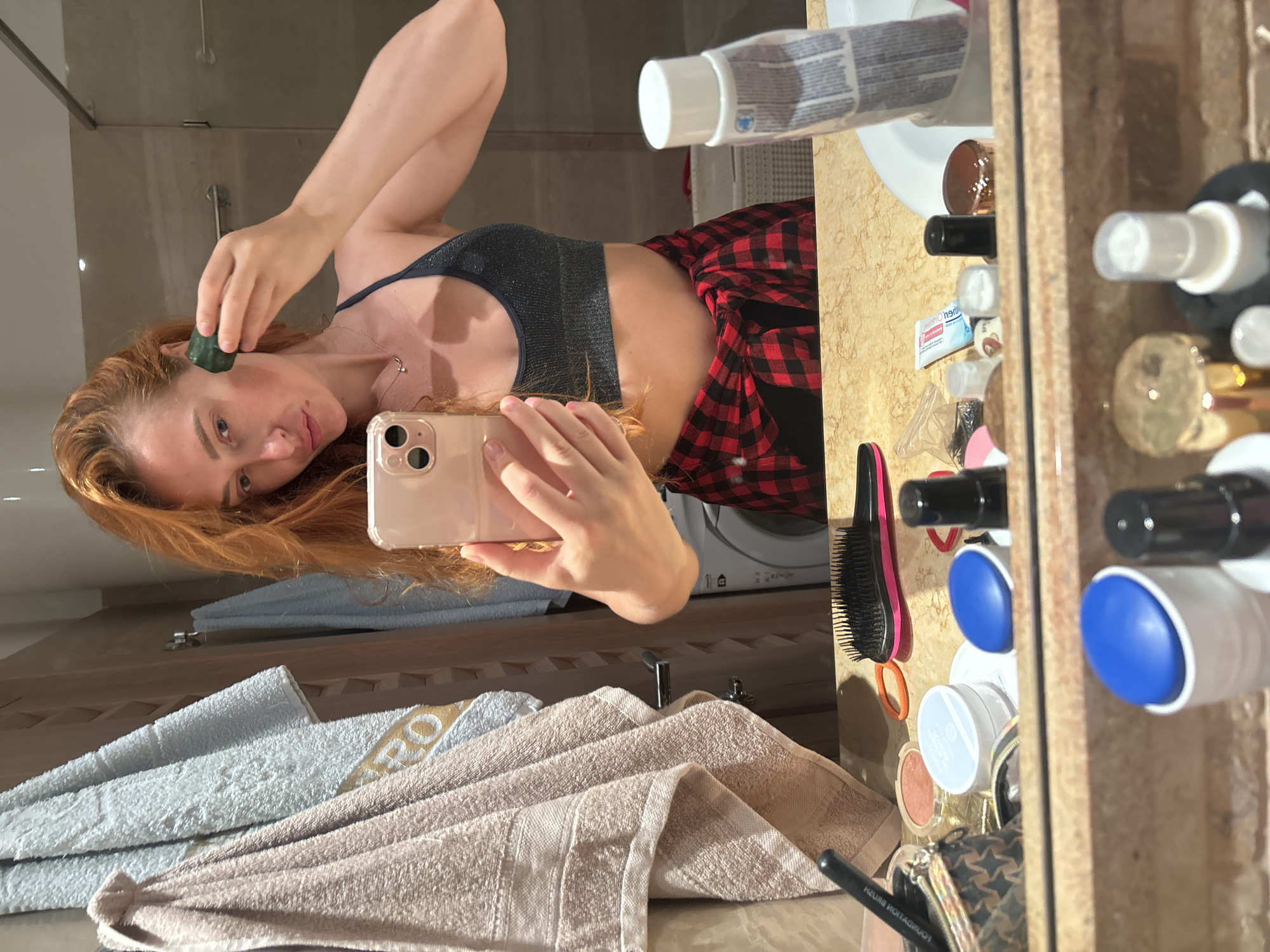 In a bathroom in Budva, Montenegro, a woman with auburn hair, wearing a dark sparkly sports bra and a red and black plaid shirt tied around her waist, stands in front of a mirror. She is holding a pink smartphone in her left hand, taking a selfie, and in her right hand, she is holding a dark green facial roller against her cheek. The mirror reflects a cluttered countertop with various cosmetic products, including makeup, brushes, and containers. Behind her, towels hang on a rack, and a washing machine is visible in the background. The lighting suggests it is daytime.