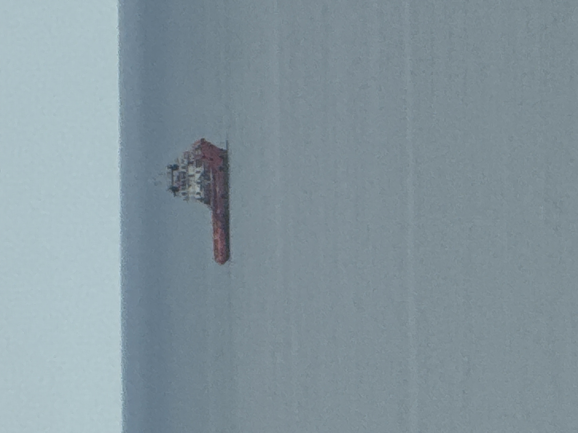 An aerial view shows a solitary red and white ship sailing on calm, gray water under an overcast sky. The ship, appearing small in the vast expanse of the ocean, is oriented towards the right of the frame. The horizon line is visible, separating the water from the sky. The image does not provide any context or indication of a specific location within the city of Kholmsk, Russia.