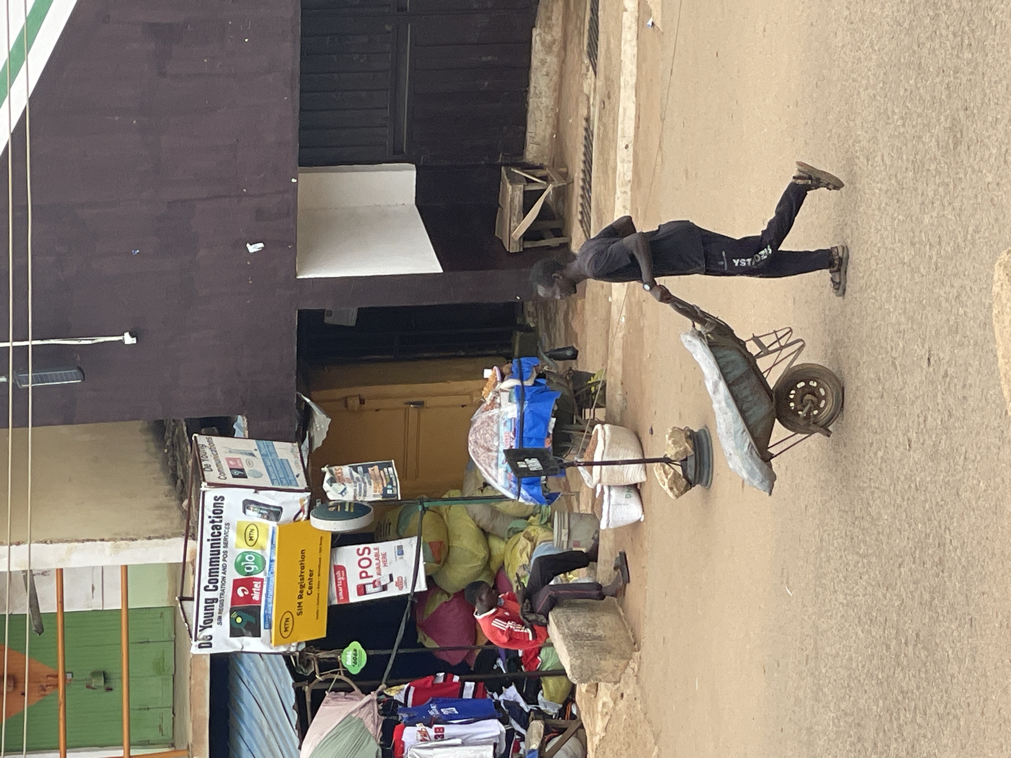 The image depicts an outdoor street scene in an unknown city. The setting appears to be a commercial area with buildings and informal market stalls.
In the left mid-ground, a white signboard for "De Young Communications" is prominently displayed, offering "SIM REGISTRATION AND POS SERVICES" for network providers including Airtel, Glo, and MTN. A yellow sign below it specifies "MTN SIM Registration Center," and another sign below reads "smartcash POS AVAILABLE HERE." Beneath a corrugated metal awning, a male individual wearing a red and white long-sleeved shirt and dark pants is seated on a concrete block, looking down at a mobile device in his hands. He is wearing flip-flops. Behind him are several large, bulging bags, some yellow and some pink, along with various hanging clothes, including sports jerseys. To his right, a small table covered with a blue patterned cloth holds items under a clear dome or cover. On the ground nearby, two white sacks containing granular material are visible.
The background on the left features a building with a green door and an orange railing on an upper level. The building on the right is a dark brown structure with a distinct white and green diagonal stripe pattern near the top right corner. A wooden stool or small table is positioned against this wall.
In the right foreground and mid-ground, another adult male, wearing a black t-shirt and black track pants (with white lettering on the left leg, possibly "YST OSA"), is pushing a dark-colored wheelbarrow containing a light-colored, tarp-like material. He is moving from left to right across the frame and is wearing sandals. The ground surface is an unpaved or dirt road. A metal grate is visible on the ground near the wall on the right.