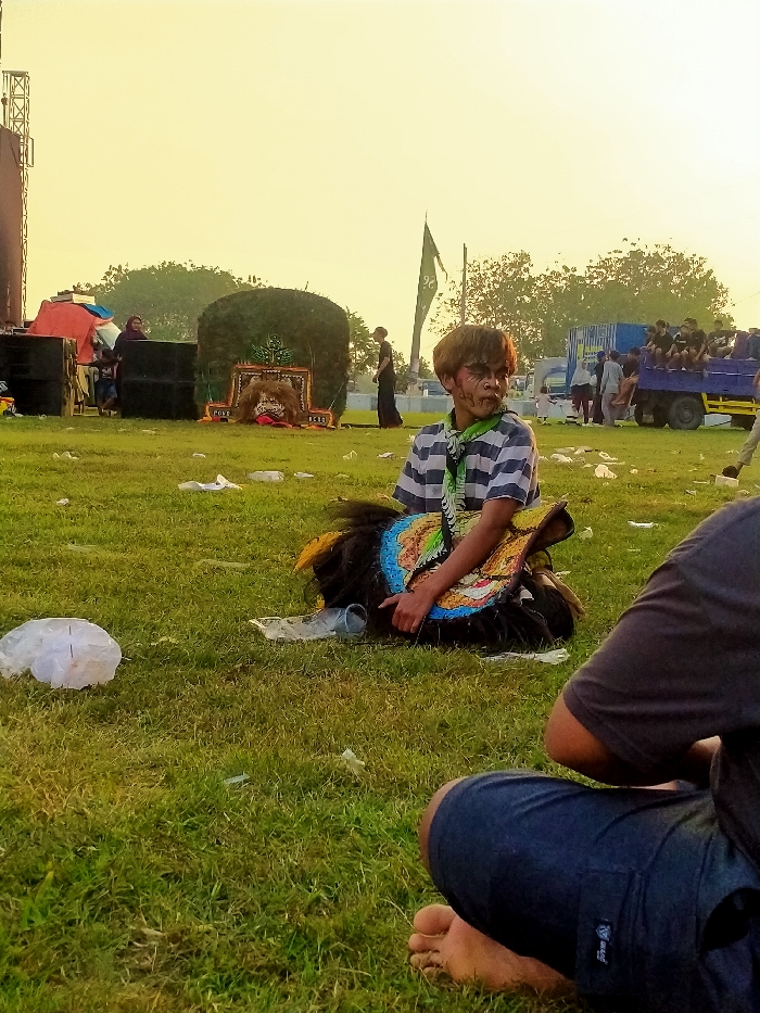 The image depicts an outdoor, grassy field scene in Glonggong, Indonesia, illuminated by warm, low-angle sunlight, indicating late afternoon or early evening. In the central mid-ground, a young male with brown hair and dark facial markings sits on the grass. He wears a striped short-sleeve shirt and a patterned neckerchief. He holds a large, elaborate costume piece with black fibrous material and colorful, intricately embroidered sections in blue, yellow, and orange, consistent with a traditional Indonesian performance prop like a Reog or Kuda Lumping element. His gaze is directed towards the right.
In the immediate foreground, the back of a person wearing dark shorts and a dark t-shirt is visible, seated on the grass with bare feet. The ground throughout the scene is green grass, sparsely covered with scattered white plastic bags and clear plastic bottles.
In the background to the left, black sound system speakers are stacked near metal scaffolding, possibly part of a temporary stage structure. A red tarp or tent is partially visible behind the speakers. Two individuals stand near this equipment. In the central background, a large, dark green, dome-like structure, heavily decorated with what appears to be foliage or fibers, is present. A stylized mask or face is visible at its base, along with a banner displaying "PONDOK PESANTREN." Further to the right, a dark-colored flag bearing a light-colored symbol (resembling '26') flies from a flagpole. On the far right, a blue flatbed truck is parked, with multiple individuals, primarily young males, seated on its bed. Other people are standing and moving in the vicinity of the truck and the background. Trees are visible on the distant horizon.