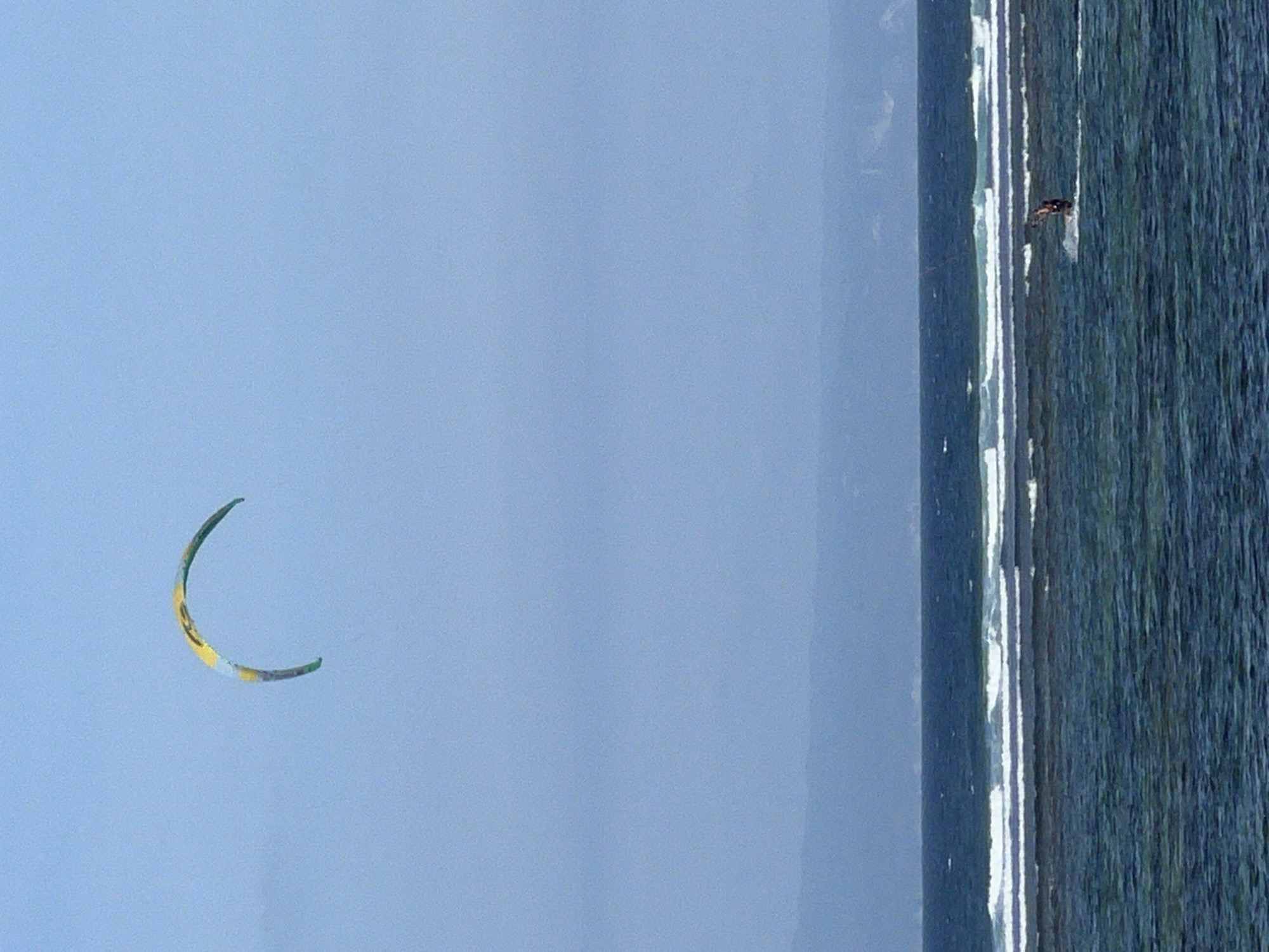 The image depicts a kitesurfer in action against a bright blue sky and the ocean. The kitesurfer, a small, dark figure, is seen on the water near the shoreline. The ocean is a deep blue-green, with whitecaps indicating choppy conditions. A surf line is visible along the coast, suggesting a beach or a shallow area.
The kitesurfer is actively engaged in the sport, as indicated by their posture and the position of the kite. The kite itself is a crescent shape, predominantly green and yellow, suspended in the air above the water. It appears to be a single kite, likely used for recreational kitesurfing.
The scene is set during what appears to be daylight, given the brightness of the sky and the clear visibility. The weather seems fair, with a clear sky and perhaps a steady breeze, ideal for kitesurfing. The location context provided is Denpasar, Indonesia, which aligns with the tropical, ocean-based activity. There is no visible text in the image. The overall impression is one of a dynamic and active moment captured in a watersport setting.