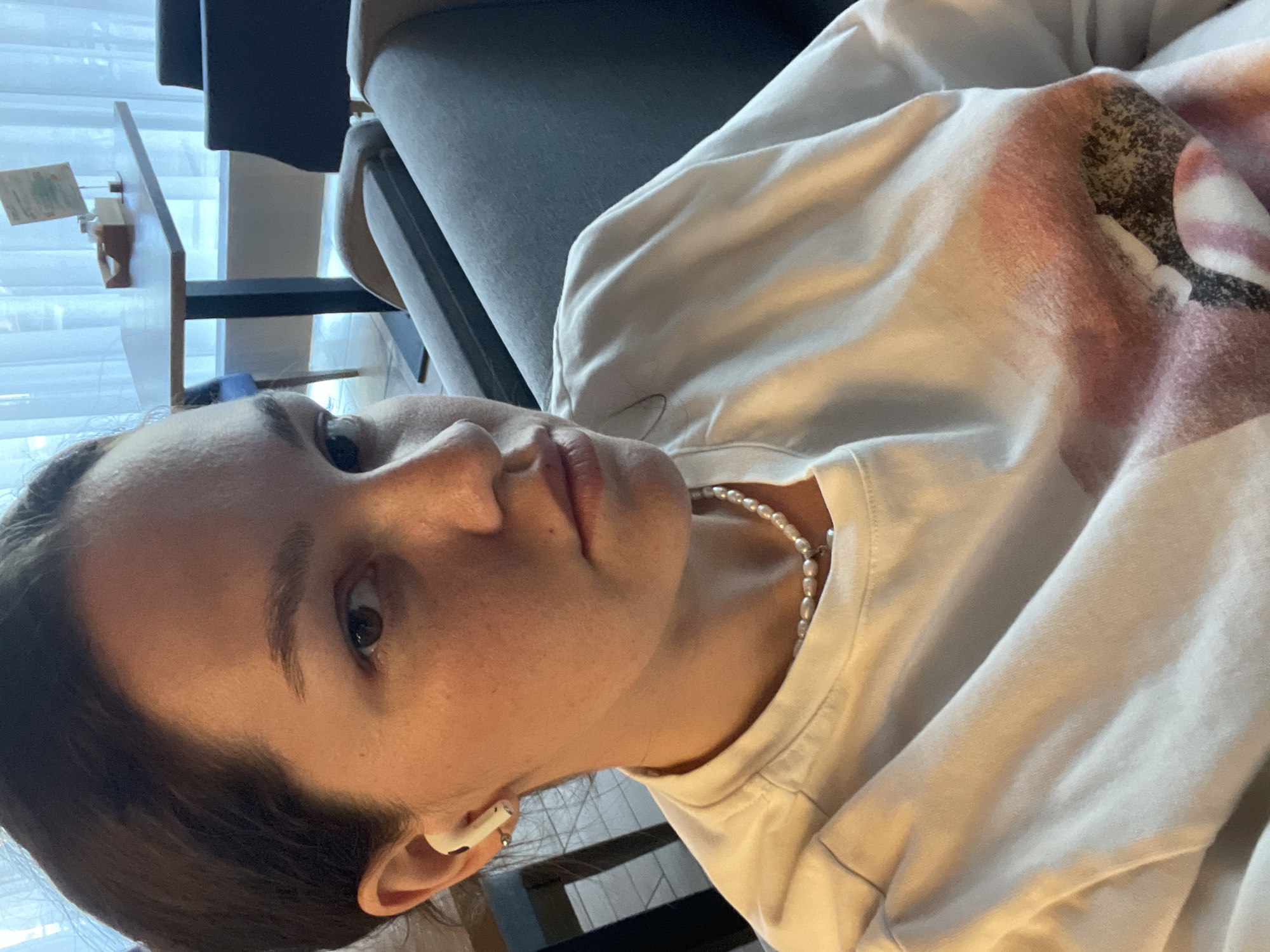 The image is a close-up, slightly tilted selfie of a woman. She is wearing a white shirt with a faded print on the sleeve, and a pearl necklace. A white earbud is visible in her left ear. She appears to be indoors, reclining against a gray couch or chair. The background is blurred, but includes a glimpse of a table and chair by a window. There is no information to suggest that this image was taken in the city of Izhevsk, Russia.