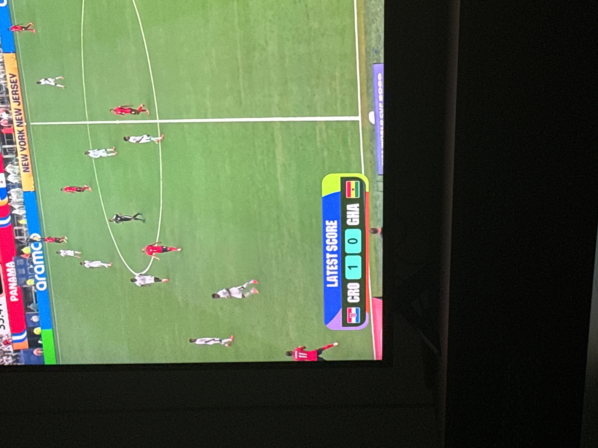 A screen displays a soccer match with a score graphic at the bottom. The score graphic shows "LATEST SCORE" with the flags of Croatia and Ghana, indicating Croatia leading Ghana 1-0. Several players in red and white uniforms are visible on the green soccer field. In the background, there are advertisements and spectators. The scene appears to be a broadcast of a soccer game. There is no indication of the city of Moscow, Russia in the image.
