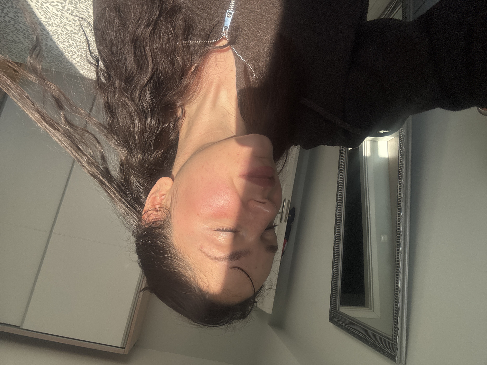 The image displays a close-up of a person, likely female, from the chest up, positioned horizontally. The primary subject has long, dark, wavy hair spread out on what appears to be a light-colored, patterned quilt or bedding. They are wearing a dark brown or black zippered top, with the zipper partially undone, revealing the neck.
Strong natural sunlight streams into the scene, creating a distinct shadow line across the person's face. The right side of their face is brightly lit, while the left side is in shadow. Their eyes are closed, and there's a noticeable reddish flush on the lit side of their cheek. Their expression appears calm or relaxed. The person's neck is visible, highlighted by the sunlight.
In the background, to the right, a large, ornate silver-framed mirror reflects part of the room, including a white door or wall. To the left, a large, light-colored wardrobe with sliding doors is partially visible. The overall setting is an indoor space, likely a bedroom, characterized by the bedding, wardrobe, and mirror. The strong, directional light suggests it is daytime, possibly morning or afternoon, with direct sunlight entering the room, indicative of sunny weather. There is no visible text in the image. The scene conveys a quiet, personal moment bathed in natural light.