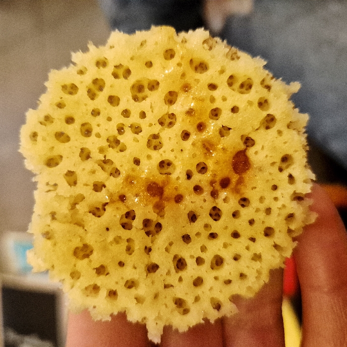 A close-up reveals a round, porous, yellow pancake-like object, likely a crumpet or honeycomb pancake. Numerous small holes cover the surface, some filled with a glistening, dark brown syrup or glaze. The edges appear slightly ragged or torn. It's being held in someone's fingers. The overall impression is of a sweet, textured treat.