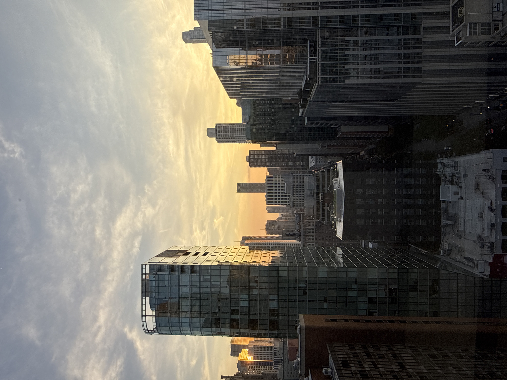 The image depicts a cityscape at dusk, likely from a high vantage point, showcasing a collection of modern skyscrapers. The setting sun casts a warm, orange glow across the sky, illuminating the clouds with hues of yellow and pink, and reflecting off the glass facades of the buildings. In the foreground, a tall, curved building with a glass exterior dominates the left side of the frame, its windows showing various shades of grey and reflecting the sky. To its right, other skyscrapers of varying heights and architectural styles are visible, some with dark glass and others with lighter facades. The scene includes numerous windows, rooftop structures, and what appear to be streets and trees far below, suggesting an urban environment. The overall impression is one of a dense, bustling city at the end of the day. The architecture and density of the buildings, combined with the specific aesthetic of the skyscrapers, strongly suggest the location is Chicago, Illinois, USA.