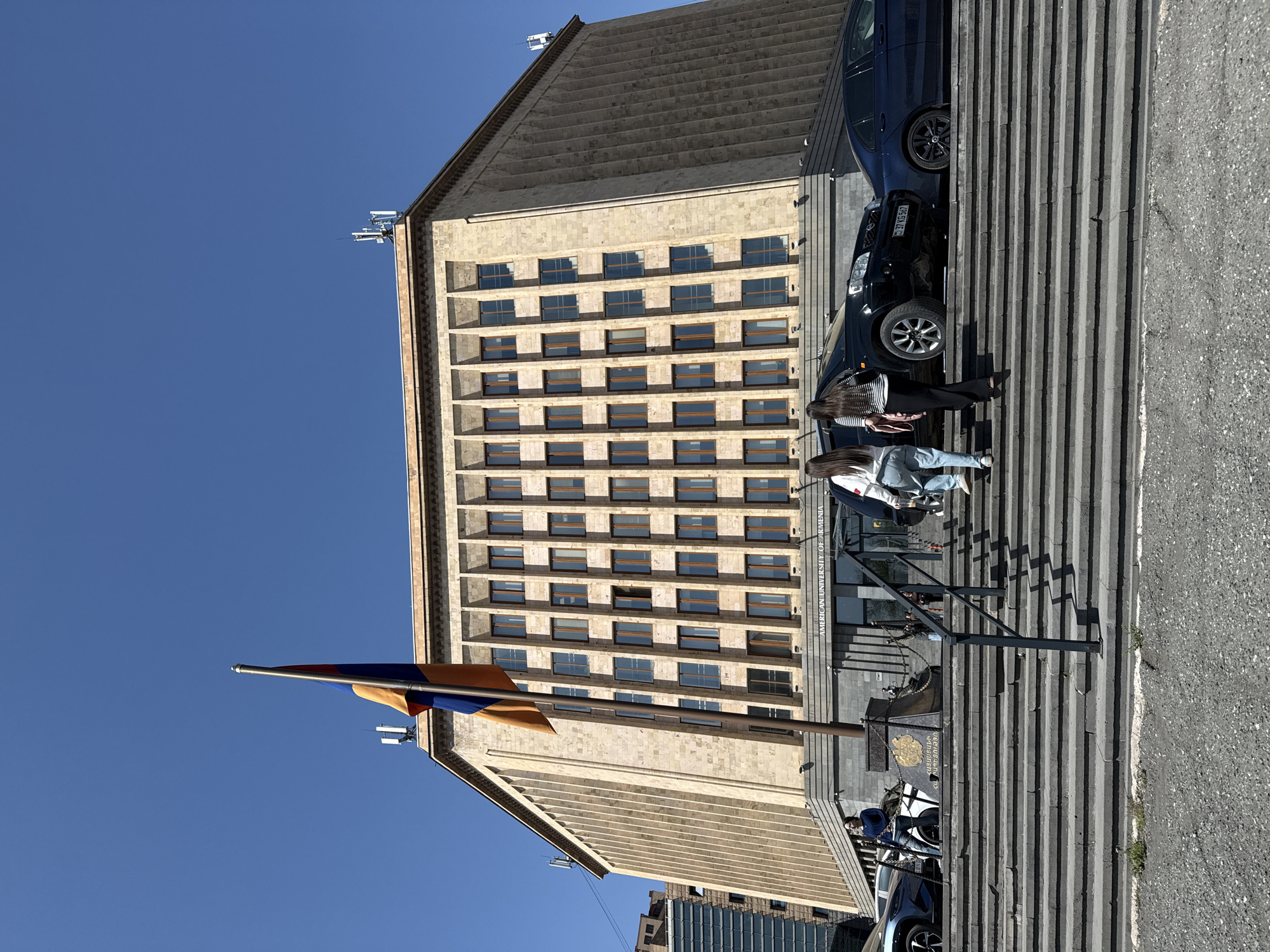 This is a daytime outdoor photograph depicting the American University of Armenia building under a clear blue sky. The image is oriented vertically (originally taken in portrait mode and rotated), showing the multi-story stone building with a wide set of steps leading up to its entrance.
The building features a light-colored, possibly tufa or granite, facade with numerous rectangular windows uniformly arranged across its multiple floors. Communication antennas or equipment are visible on the roofline on the upper right side. Above the main entrance, prominent text reads "AMERICAN UNIVERSITY OF ARMENIA," with its Armenian equivalent, "ՀԱՅԿԱԿԱՆ ԱՄԵՐԻԿՅԱՆ ՀԱՄԱԼՍԱՐԱՆ," visible below it.
On the left side of the image, the national flag of Armenia, with its horizontal red, blue, and orange stripes, flies from a flagpole in front of the building. Near the base of the flag pole and steps, a monument or plaque with text in Armenian is partially visible.
Several people are present, including two individuals with long hair walking up the wide, concrete steps on the right side of the frame. Their casual attire suggests everyday activity. To the right of the steps, two dark-colored cars are parked. The license plate "37 AG 047" is clearly visible on one of the vehicles, confirming an Armenian registration.
The clear, vibrant blue sky and the distinct shadows cast by the building and people indicate a sunny day, likely in the late morning or early afternoon. The scene is an urban setting, specifically in Yerevan, Armenia, as confirmed by the university's name, the national flag, and the vehicle's license plate.