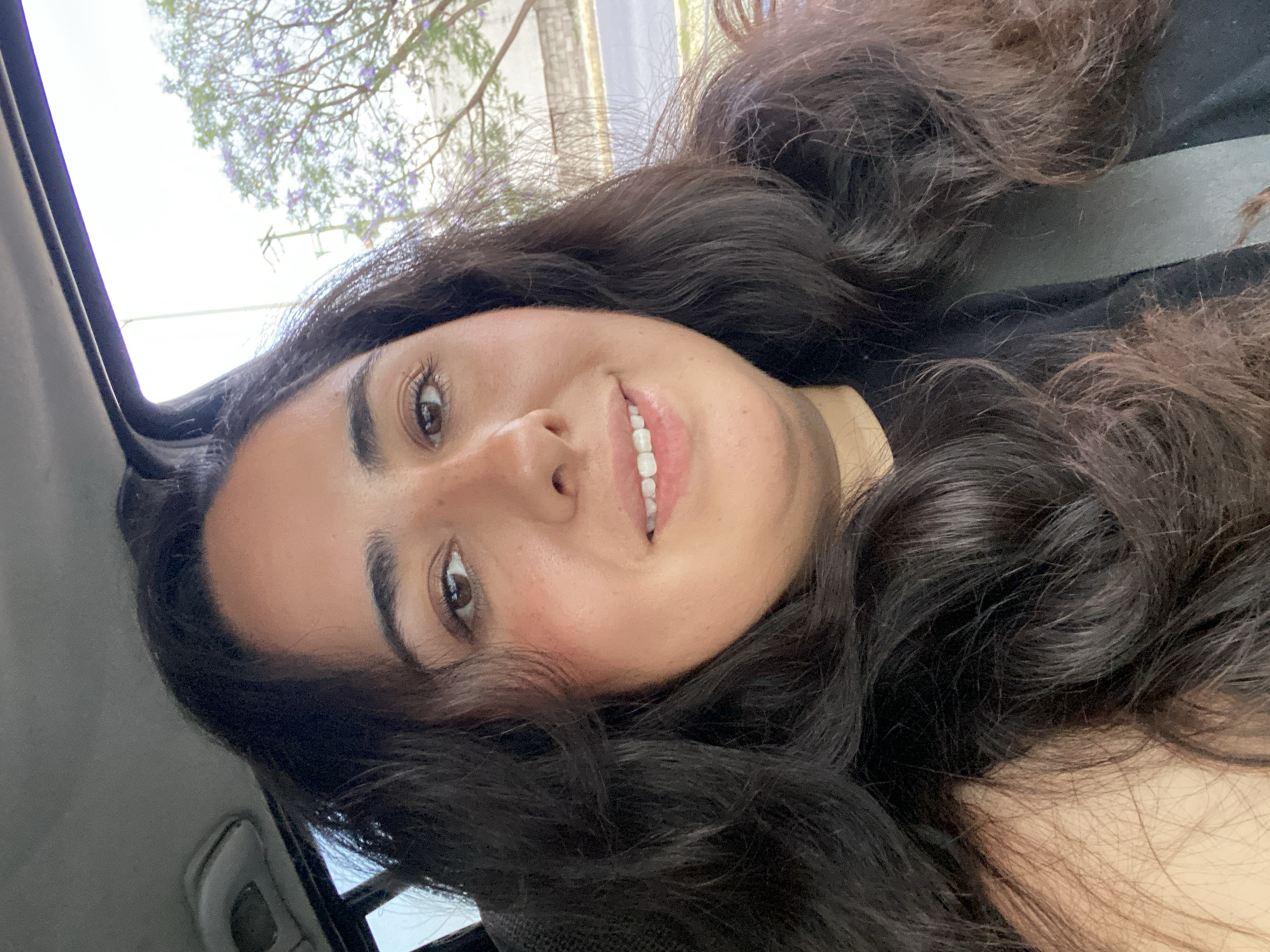 A woman is angled and cropped in a selfie, likely taken in a vehicle with an open sunroof. The woman is smiling, and her dark, wavy hair frames her face. Outside the sunroof, tree branches with purple blossoms are visible. The location is San Bernardino Tlaxcalancingo, Mexico, as inferred from the prompt. Additional details are not discernible from the provided image.