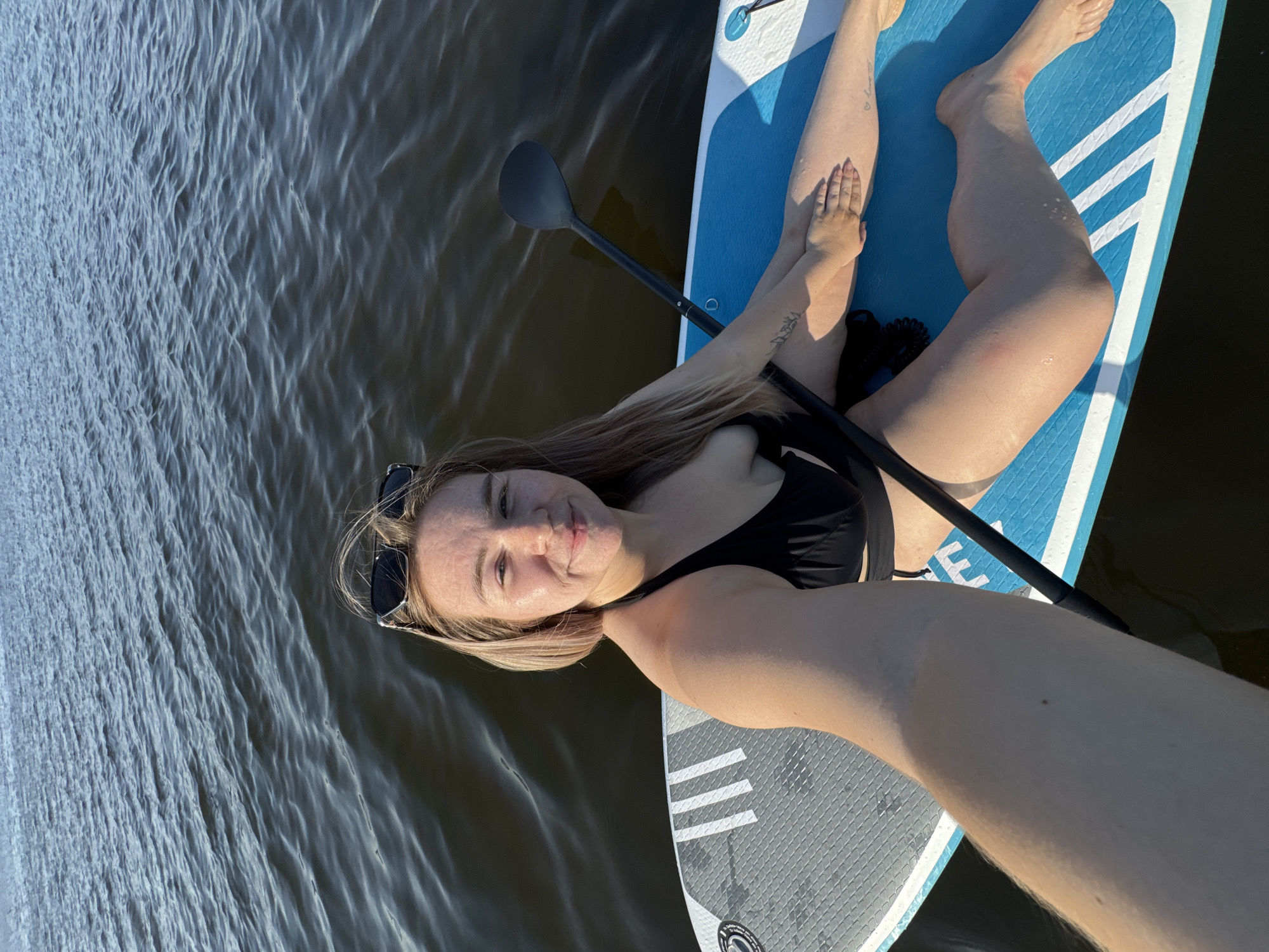 A woman is lying on a stand-up paddleboard on a body of water. She is wearing a black bikini and sunglasses. A paddle is lying next to her. The water is slightly choppy. The location is likely near Chaikovsky, Russia.