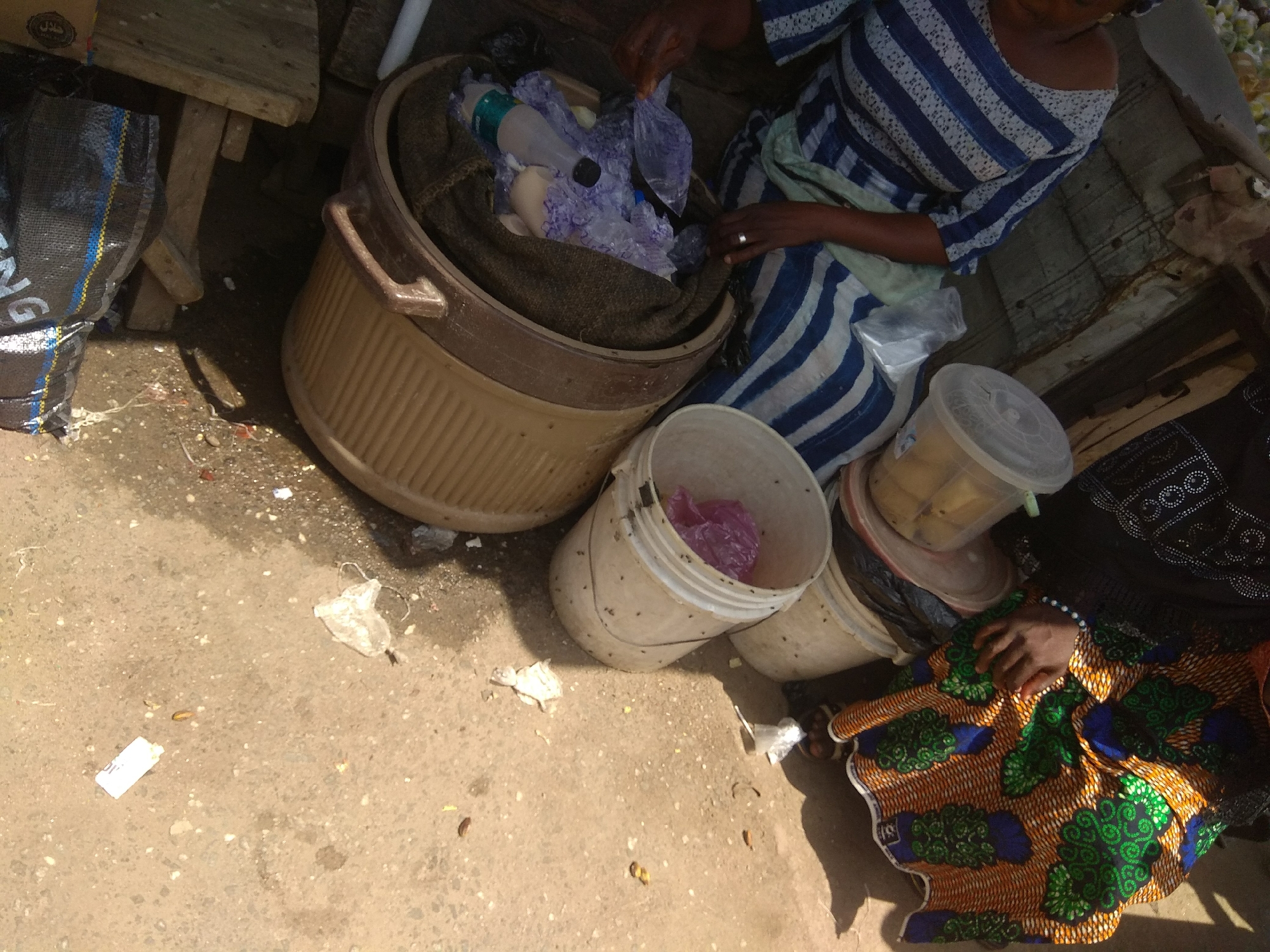 This image depicts an outdoor scene in Jalingo, Nigeria, likely a market or a street vending area. The central focus is a large, light-colored plastic tub containing a dark cloth lining and various items, including plastic bottles and purple-wrapped objects. A person wearing a blue and white striped dress is visible, with their hands reaching into the tub, suggesting they are arranging or sorting the contents.
Nearby, there are several other containers. A white bucket holds a purple plastic bag. Another white bucket is partially visible. To the left, a large, black and blue woven bag stands upright. The ground is a mixture of sand and dirt, with scattered debris, including pieces of white plastic and what appear to be dried leaves or small pieces of food.
The lighting suggests it is daytime, possibly with bright sunlight casting shadows. The overall setting appears somewhat informal and potentially rustic. The presence of multiple containers and a person actively engaged with the goods implies a commercial or work-related activity. The image is shot from a slightly elevated perspective, looking down on the scene. There is no visible text in the image.