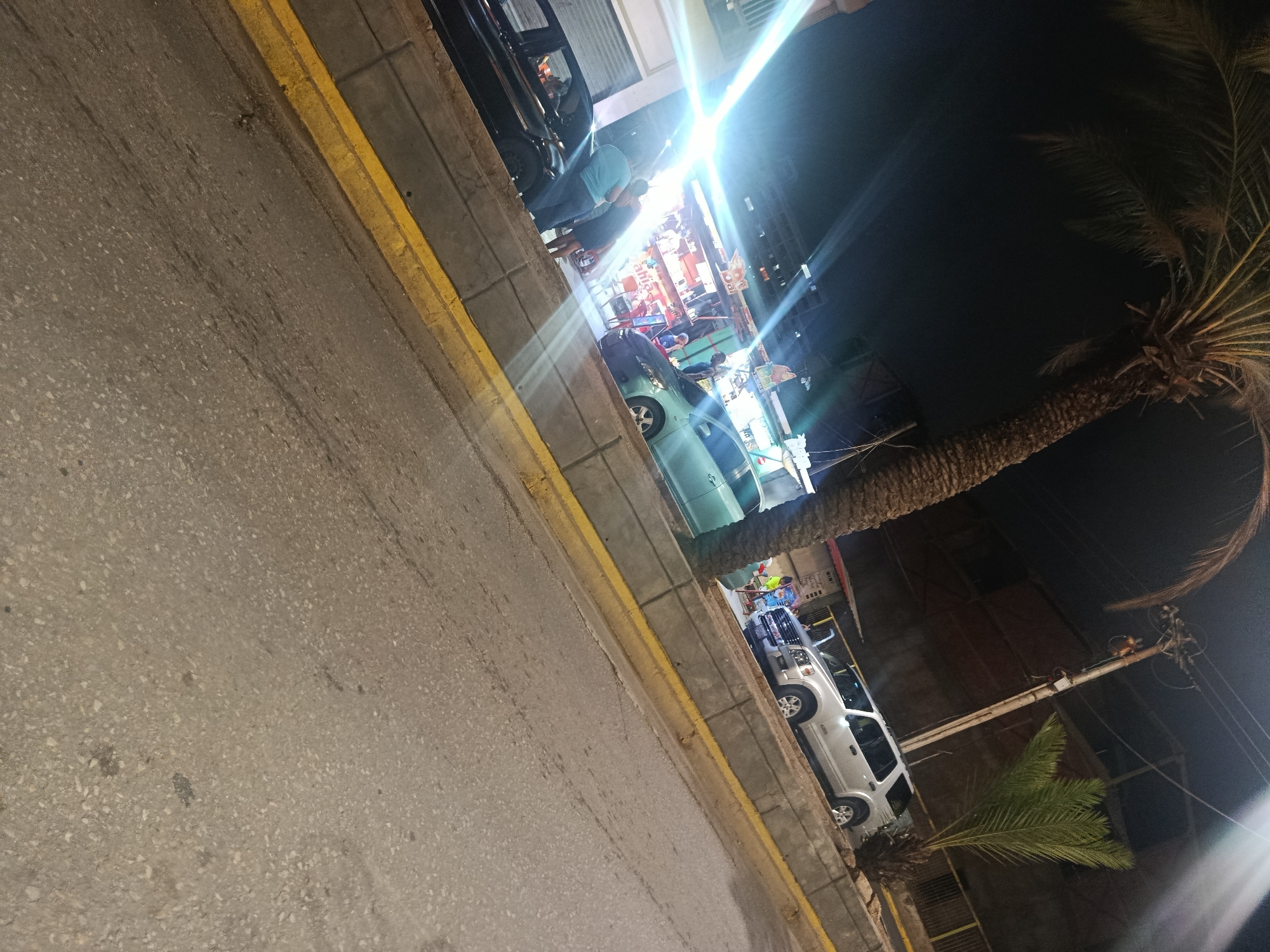 The image depicts a night-time urban street scene in Juan Griego, Venezuela, captured with a rotational tilt of approximately 45 degrees clockwise. The foreground displays an asphalt road surface bordered by a yellow-painted concrete curb and an adjacent concrete sidewalk. Along the curb, multiple vehicles are parked: a light blue compact car is visible on the left side, and a light-colored SUV, likely silver or white, is positioned further right. Behind these vehicles, a row of commercial establishments, resembling shops or stalls, are brightly illuminated by artificial light sources, generating significant lens flares across the image. Several individuals are present in this area; specifically, two individuals, one in a light blue shirt and another in a darker shirt, appear to be interacting near a dark-colored vehicle on the left. The background features a dark night sky. A large palm tree with a thick trunk and visible fronds dominates the right side of the frame. A utility pole with overhead wires is also visible on the right, behind the SUV. A second, smaller palm tree is partially visible lower on the right. No specific identifying street names or landmarks are discernible.
