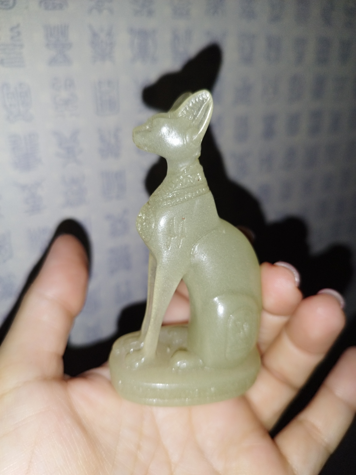 The image shows a person holding a small, pale green, translucent figurine of a seated cat, possibly made of jade or a similar stone, in their left palm. The cat figurine has large ears, a slender body, and is adorned with a decorative collar and some engraved details on its flank. The background is a light-colored wall with a repeating pattern of blue, somewhat blurry Chinese characters. A shadow of the figurine and the hand is cast on this wall to the right. The person's fingers are visible, with light pink nail polish on the nails of the right hand. The scene is lit from the side, creating highlights on the figurine and casting shadows. The image appears to be a close-up, indoor photograph. There is no indication of any specific location in Saint Petersburg, Russia.