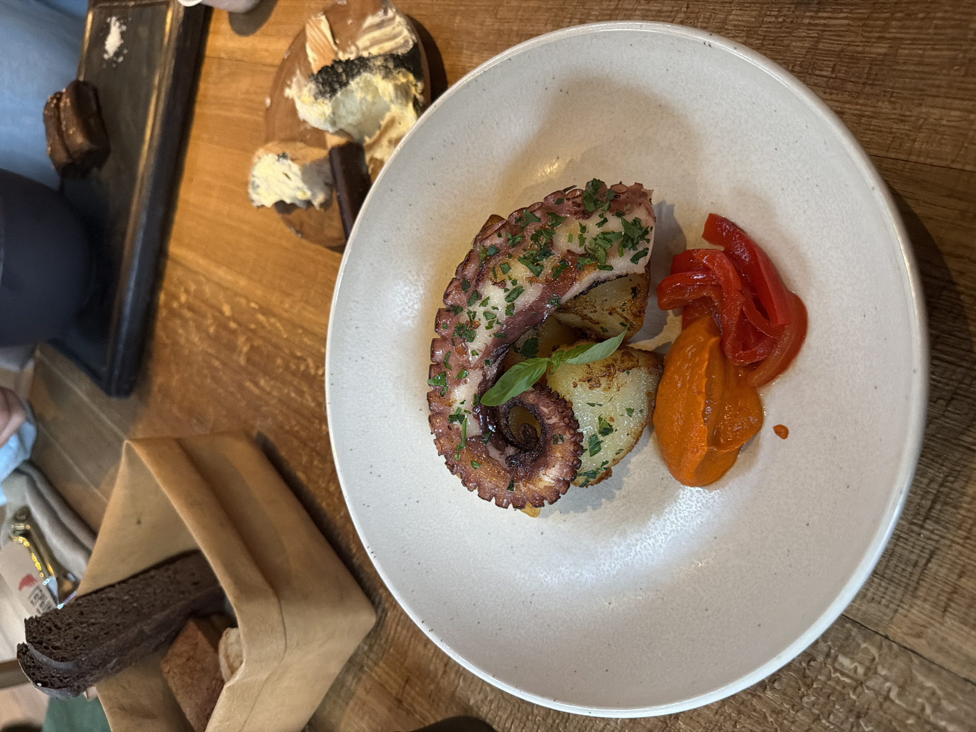 The image depicts a meal at a restaurant. A white plate dominates the scene, featuring grilled octopus with herbs, potatoes, red pepper slices, and a red sauce. Adjacent to the plate, there's bread served in a tan paper bag and a board with cheese and dark squares (possibly chocolate). The table has a wooden surface. The location is likely Moscow, Russia, given the file name, but there is no specific information within the image itself to confirm this. People's hands are partially visible, suggesting the meal is being shared or enjoyed by multiple individuals.