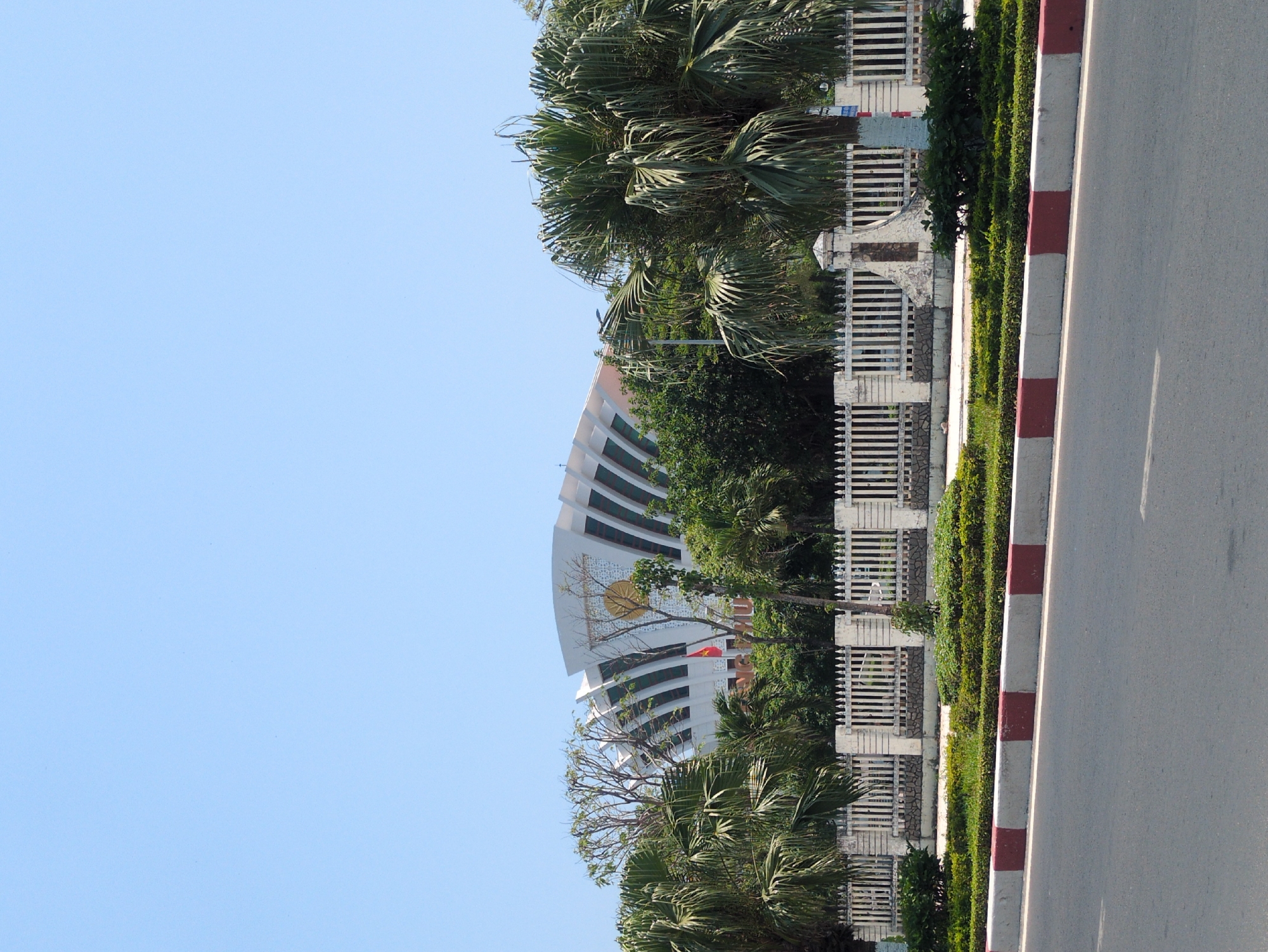The image shows a building with a curved facade, possibly a government building, located behind a fence and some greenery along a road. The building is white with visible horizontal levels. A golden emblem is affixed to the facade. A Vietnamese flag is visible on the building. The environment suggests a city location, likely Tuy Hòa, Vietnam. The road has a red and white curb. Palm trees are positioned near the fence. The sky is blue.