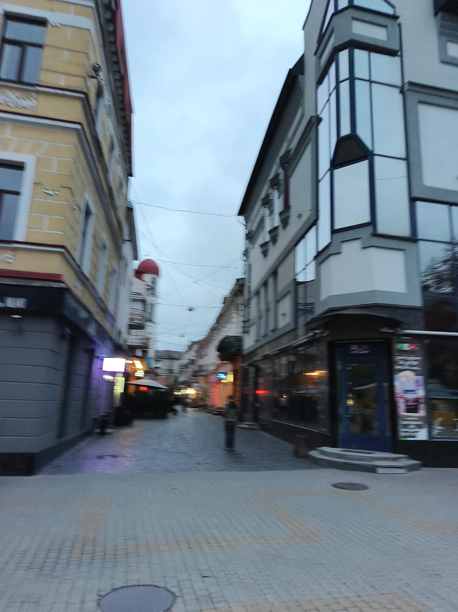 The image depicts a street scene, possibly in Ternopil, Ukraine, on an overcast day. The perspective is low and angled, suggesting the photo was taken from a moving vehicle. A paved street with textured paving stones runs through the center of the frame, leading towards a distant intersection lined with buildings. On the left, a cream-colored building with a red roofline and dark window frames is partially visible. Adjacent to it, a modern building with a grey facade and extensive glass windows dominates the right side of the street. Several storefronts are visible along the street, some with glowing signs. A few blurred figures are present in the distance, walking on the sidewalk. The sky is a pale grey, indicating muted lighting conditions.